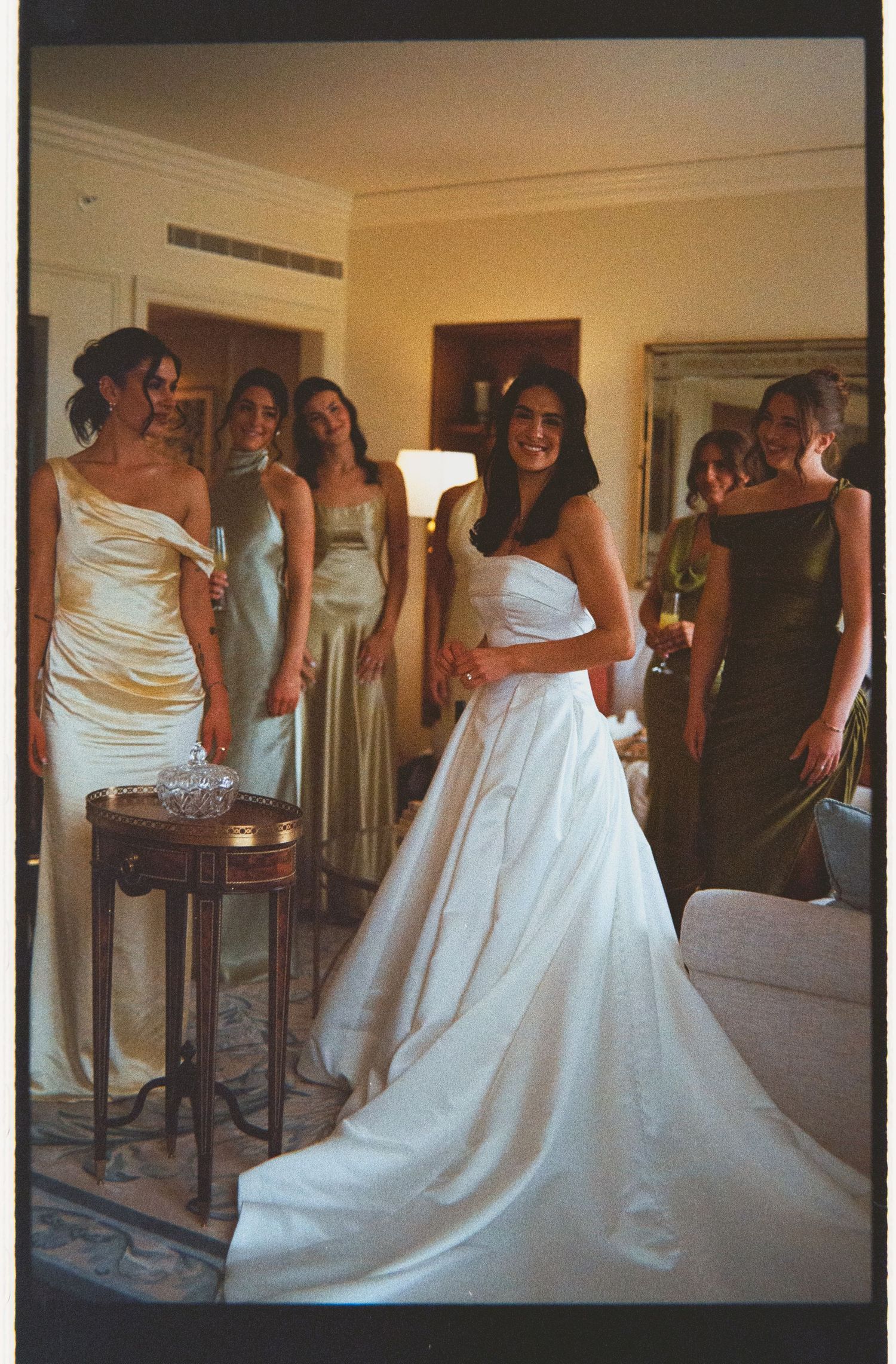 Bride in a strapless ball gown with train stands with bridesmaids in mixed neutral dresses in a hotel room.