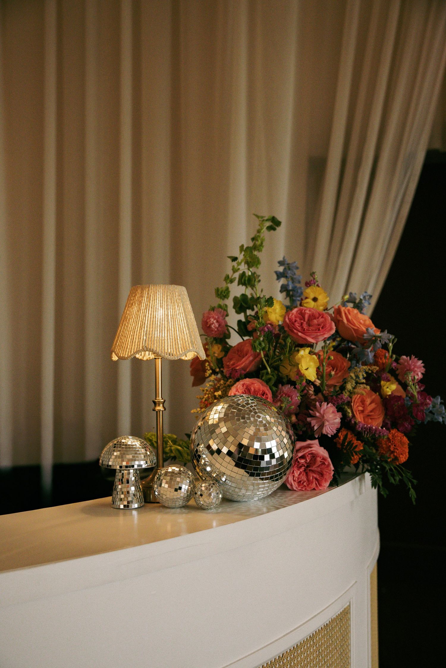 Wedding decor display with colorful floral arrangement, disco balls, and beaded lamp on white bar.