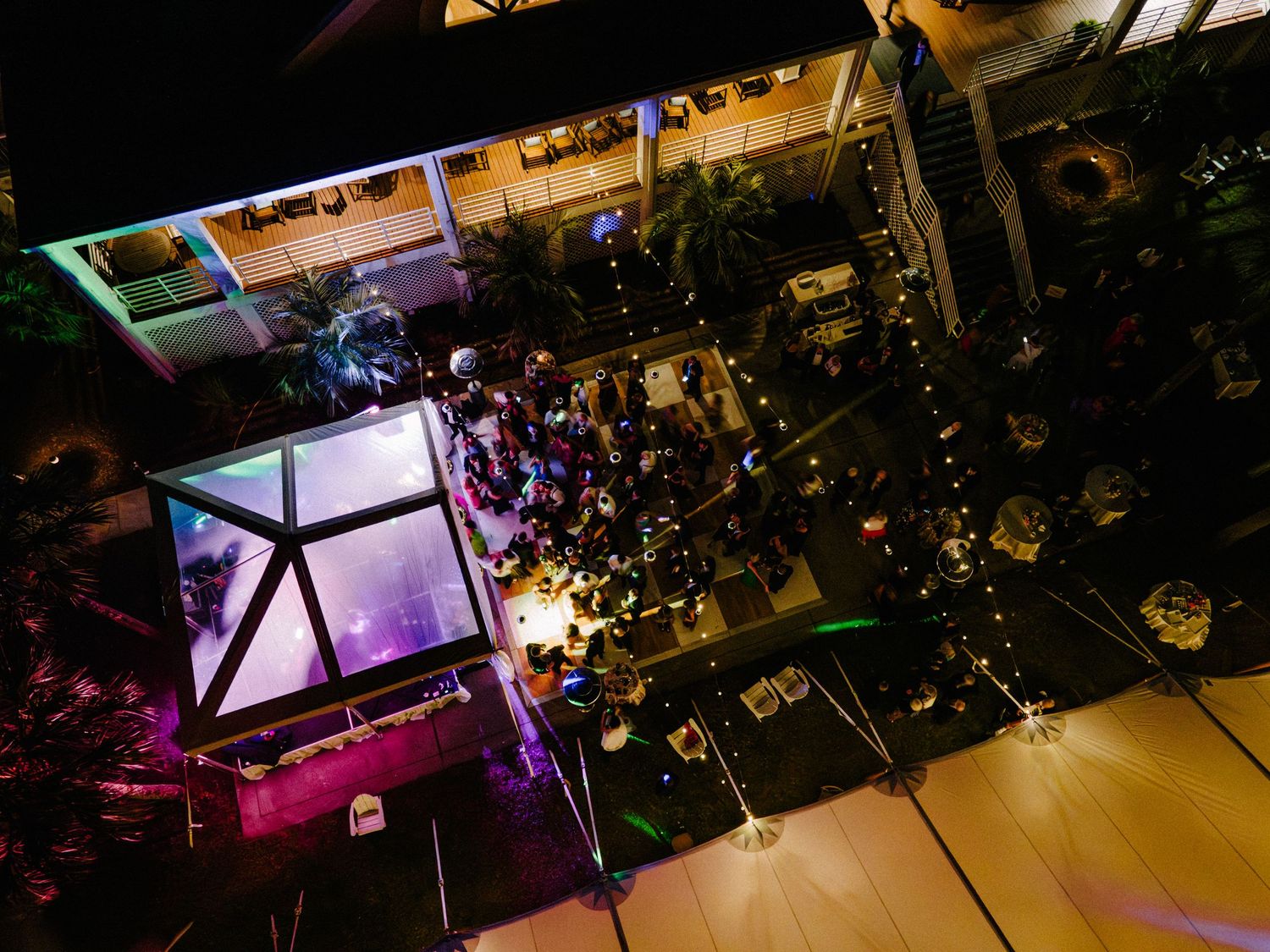 Aerial night view of an outdoor event venue with a dance floor, string lights, and crowds gathered under tropical trees.