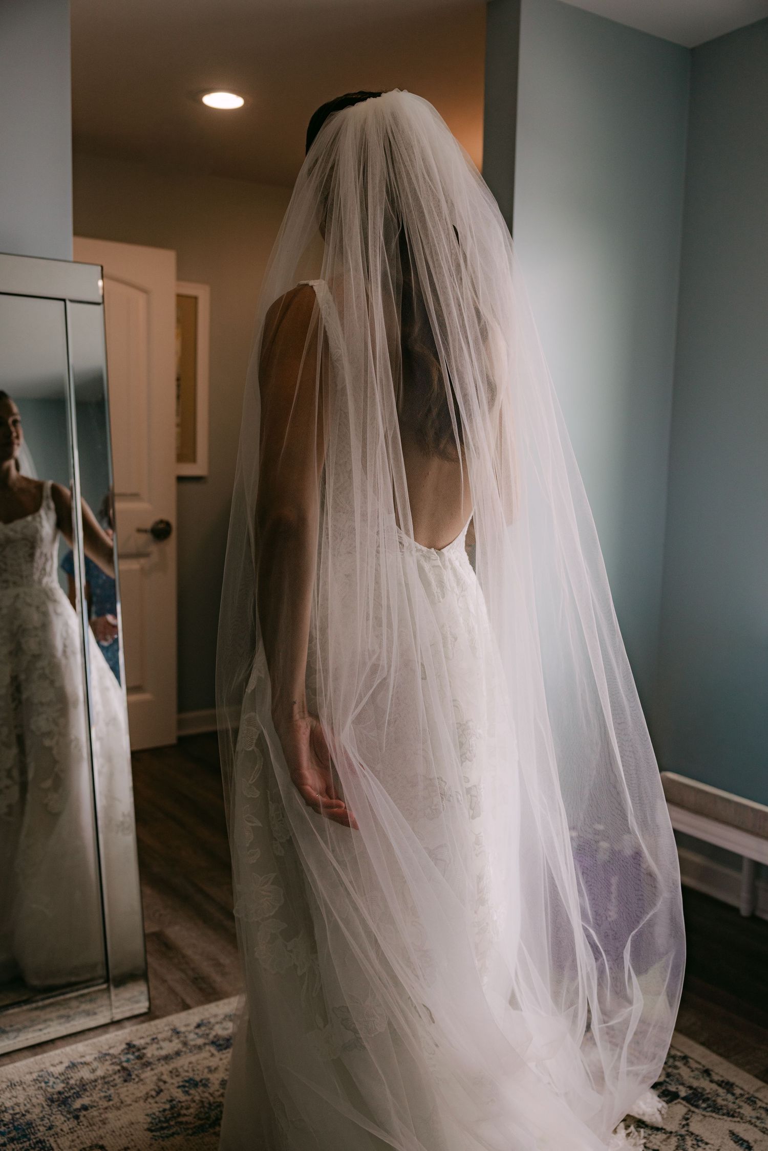 Bride seen from behind wearing lace wedding gown with long tulle veil in bridal suite.