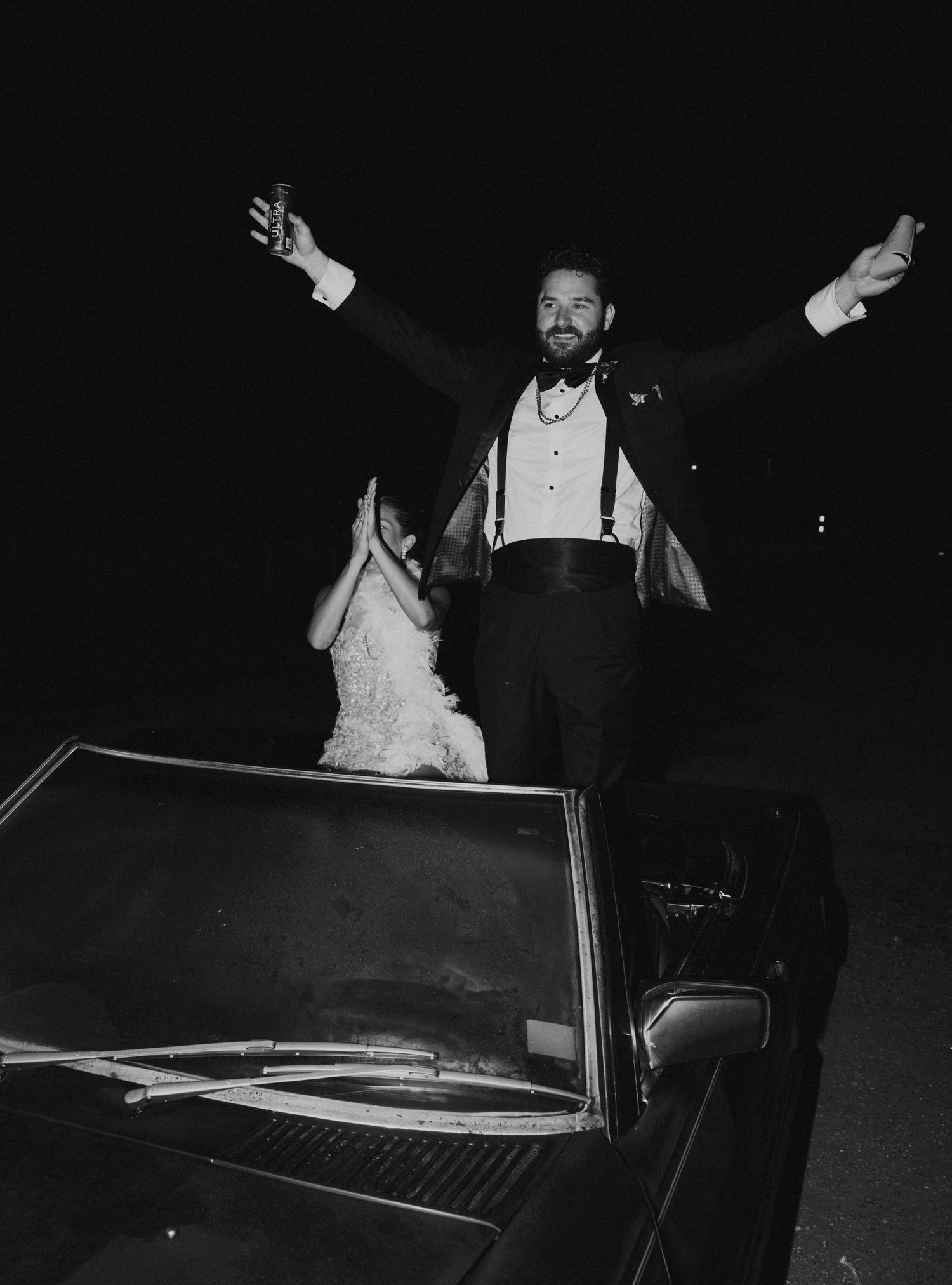 Black and white photo of groom standing in convertible car with arms raised at wedding exit.