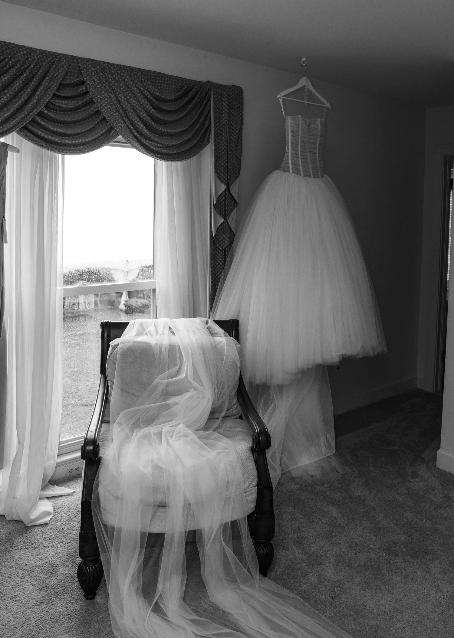 Black and white photo of a wedding dress hanging beside a chair draped with a bridal veil near a window.