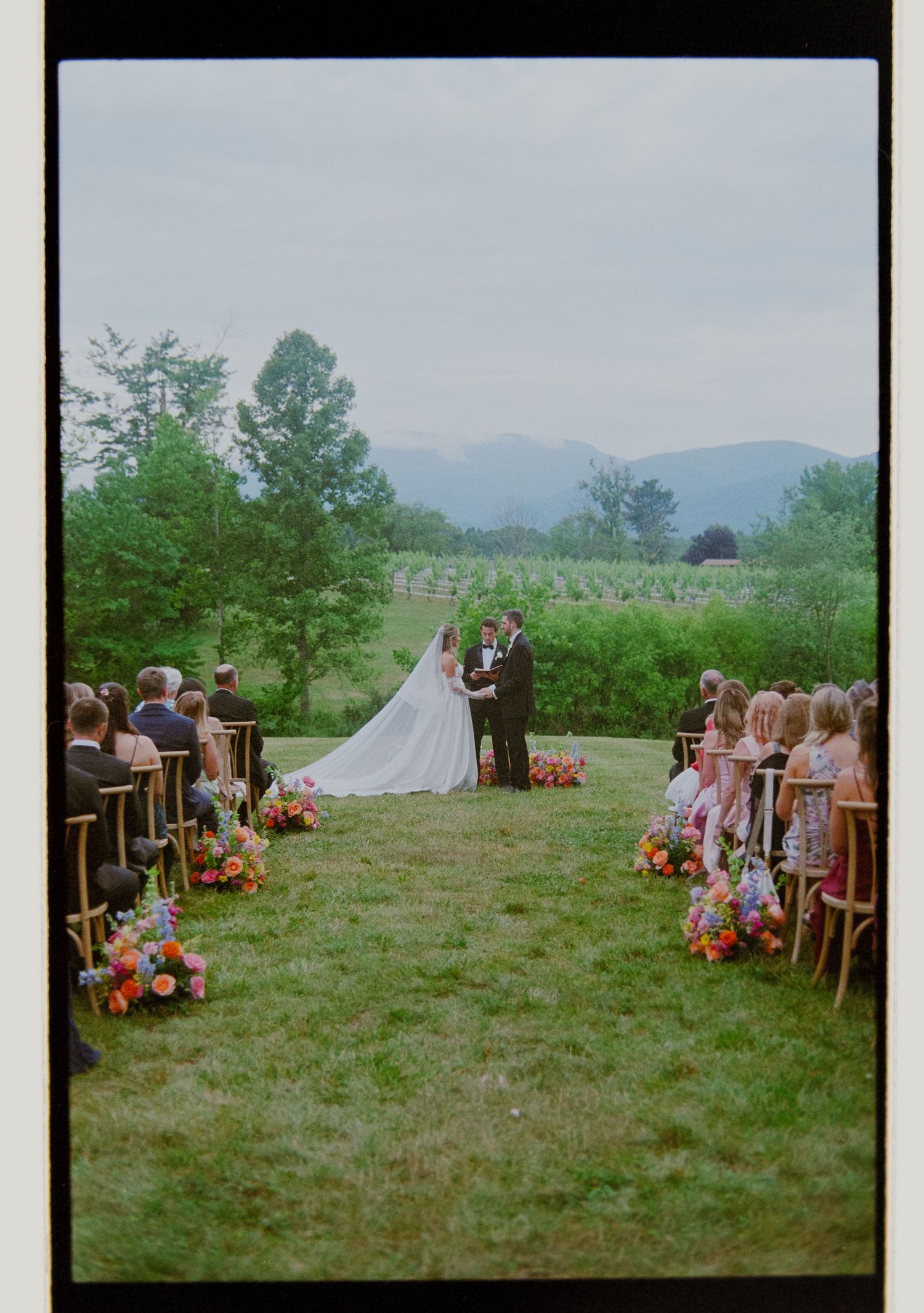 Bride and groom exchange vows at outdoor ceremony with mountain views and colorful floral aisle arrangements.