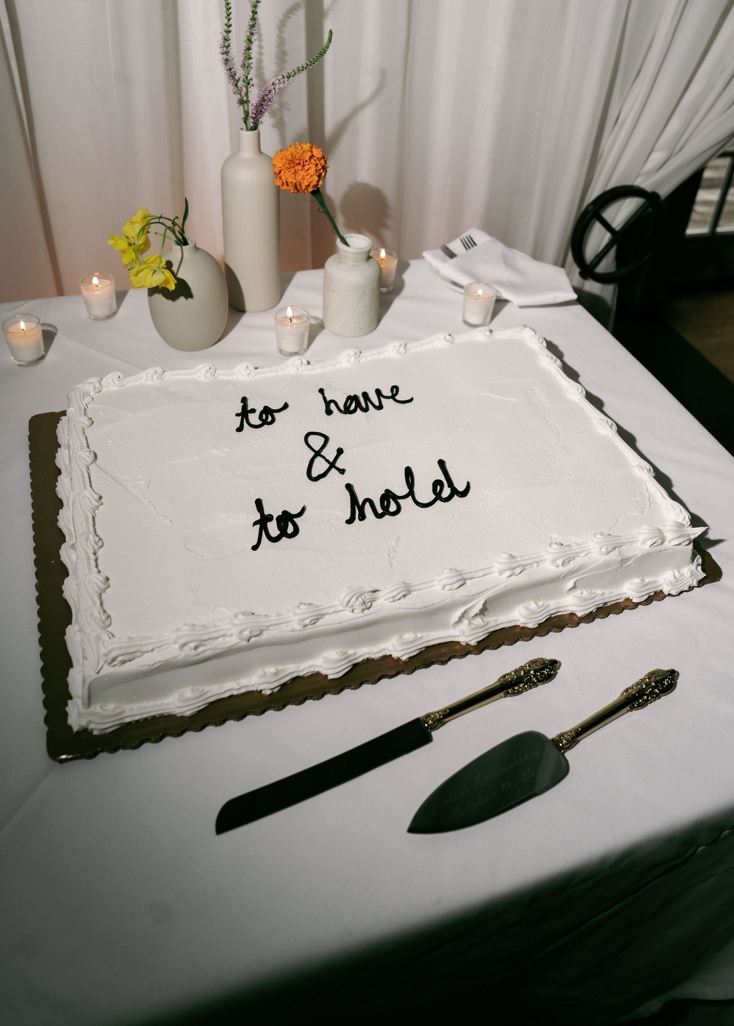 White sheet wedding cake decorated with script reading to have and to hold beside silver cake server set.
