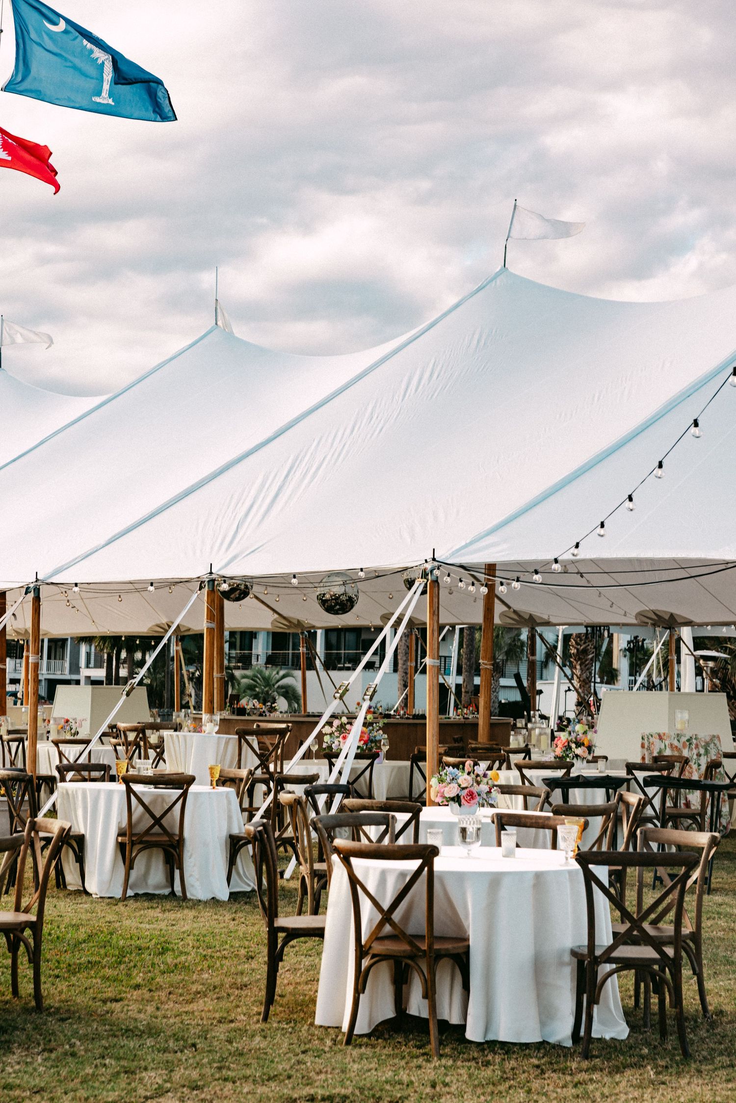 Elegant outdoor wedding reception under a large white sailcloth tent with string lights, wooden cross-back chairs, and white linen tables.