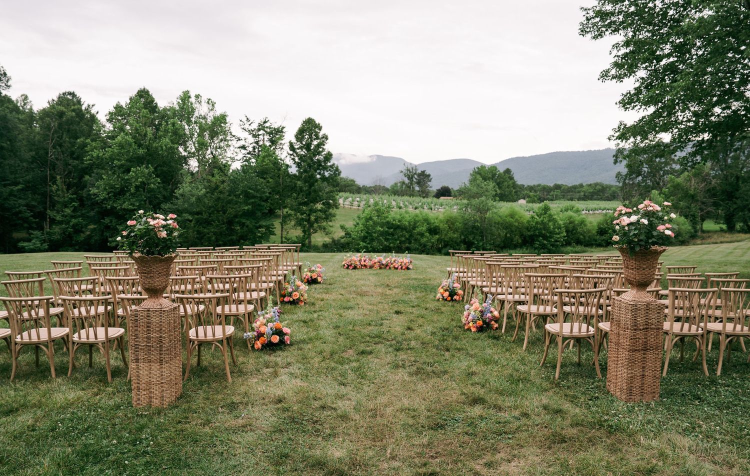 Outdoor wedding ceremony setup with wicker urns, floral arrangements, and wooden chairs on a green lawn with mountain views.