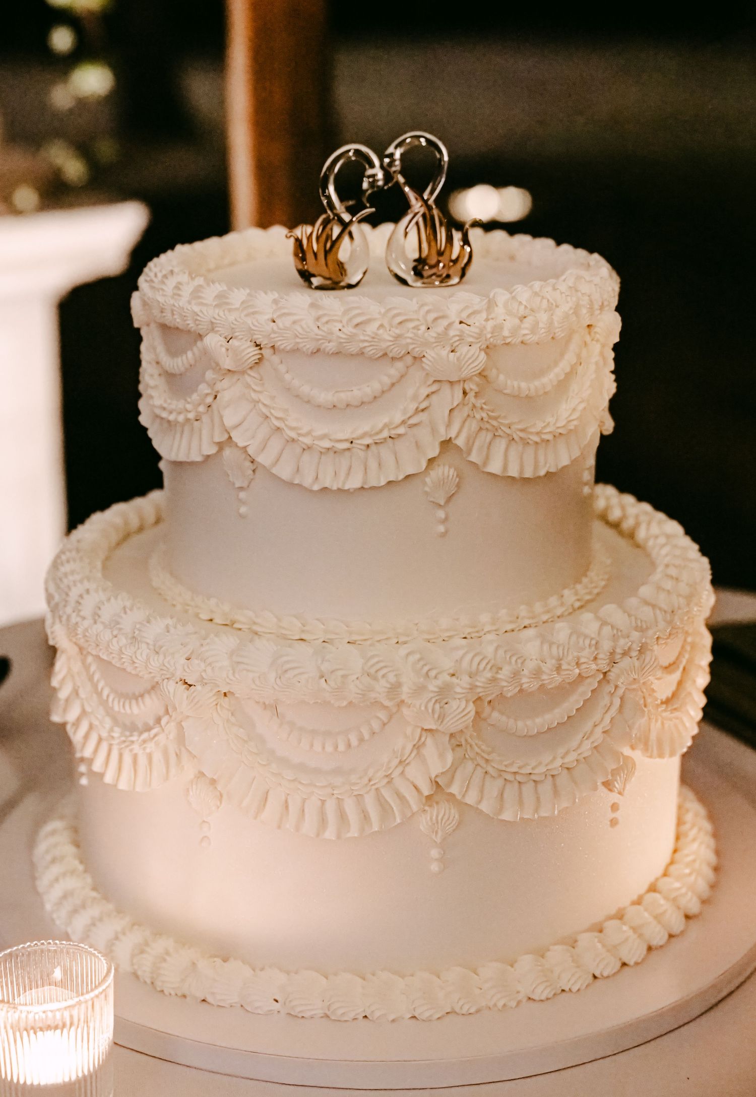 Elegant two-tier white wedding cake with intricate ruffle frosting details and golden swan cake toppers.