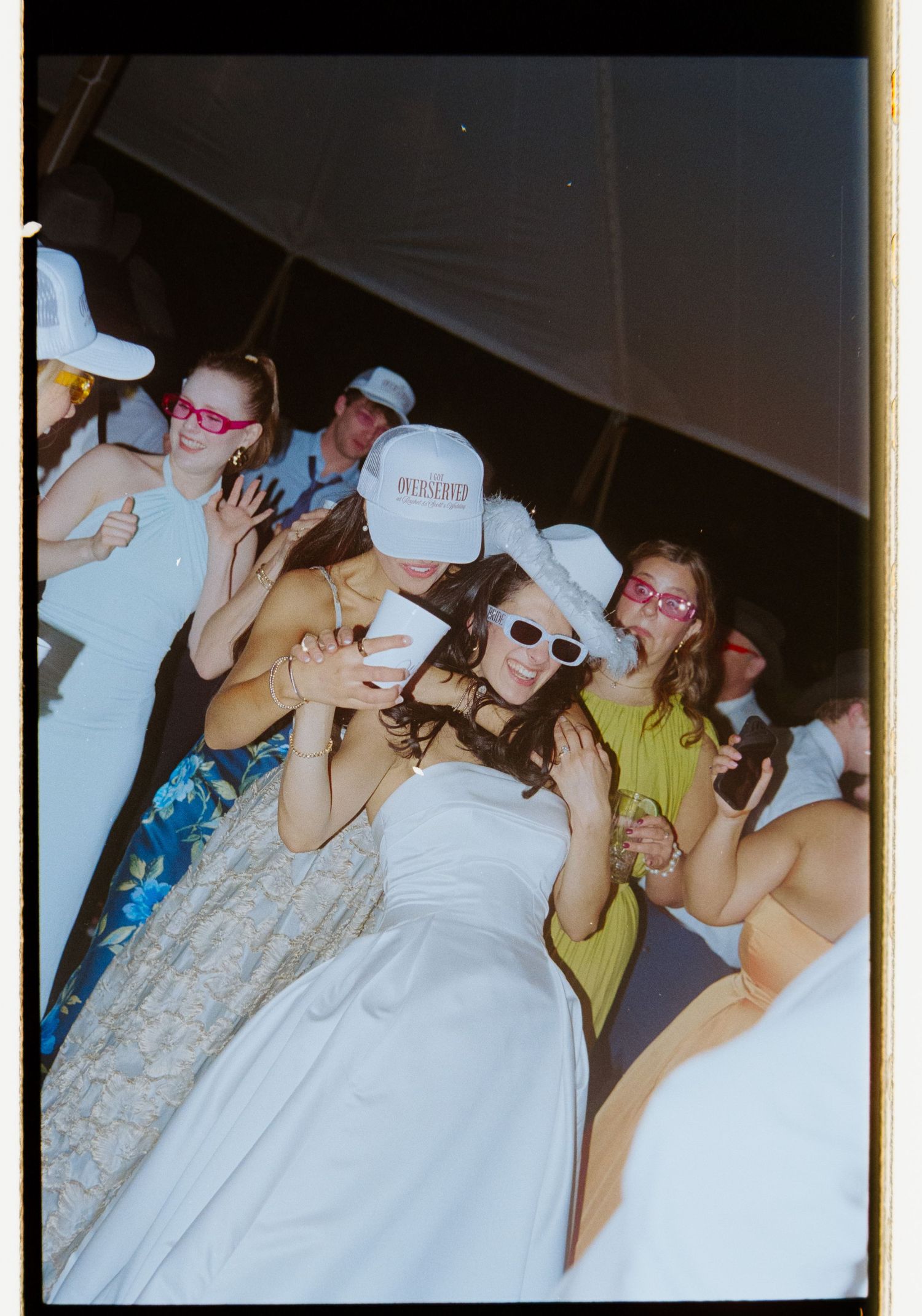 Bride dances with guests wearing white caps and sunglasses at lively wedding reception.
