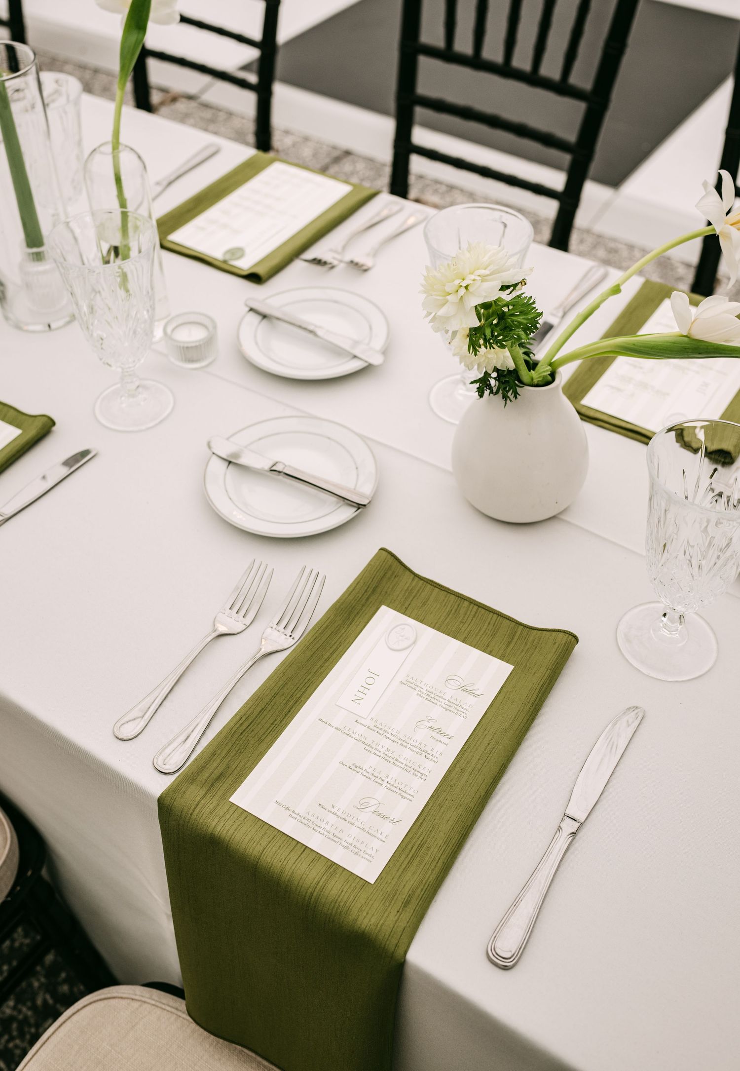 Elegant wedding reception table setting with green silk napkins, white plates, silver cutlery, and white floral centerpiece.