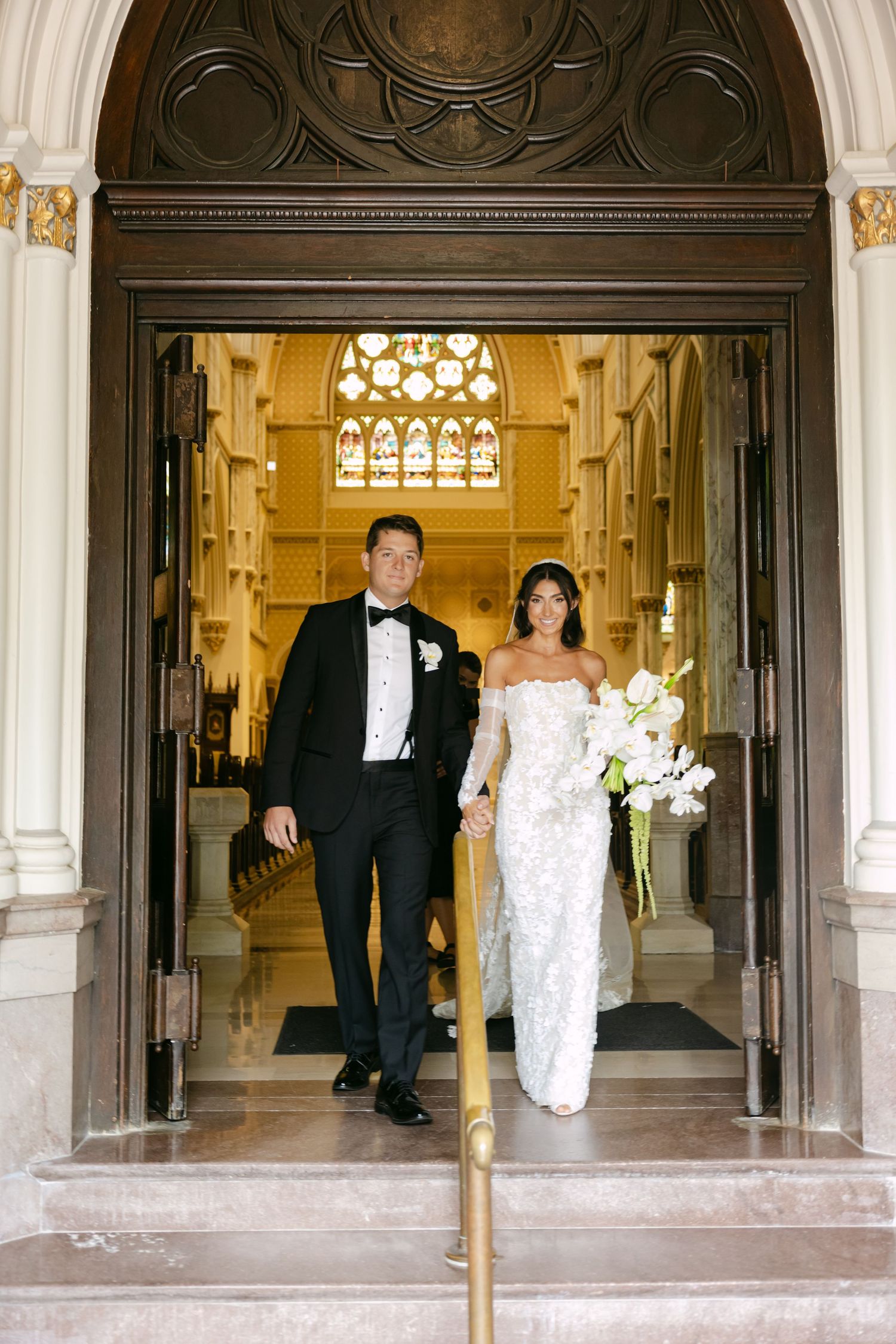 Wedding couple walks through grand church entrance with arched doorways and ornate details.