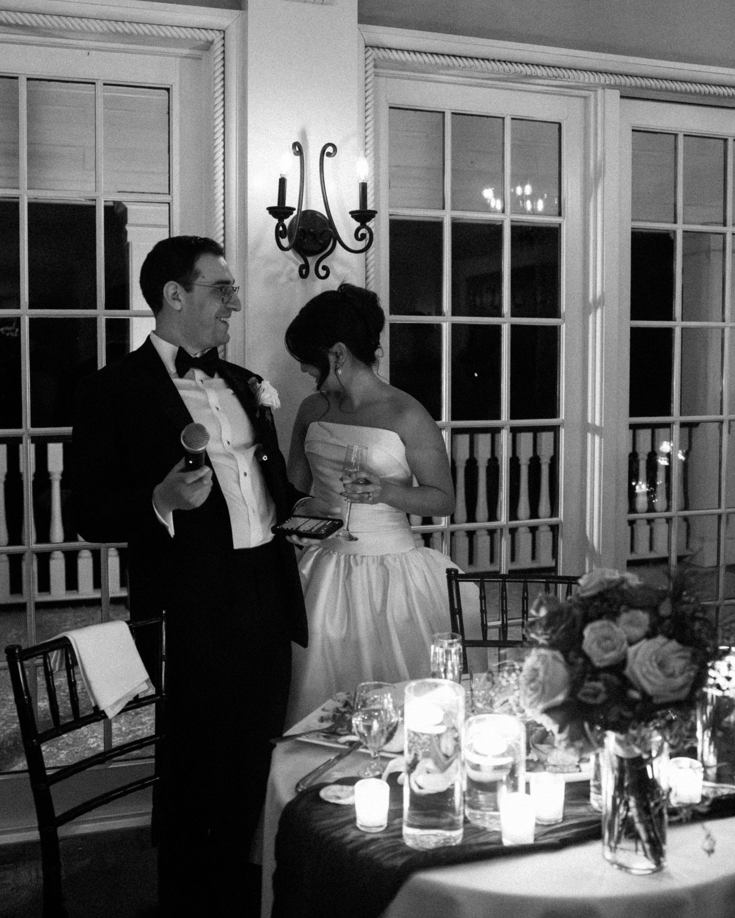 A couple in formal attire share an intimate moment at an elegant candlelit reception with floral centerpieces.