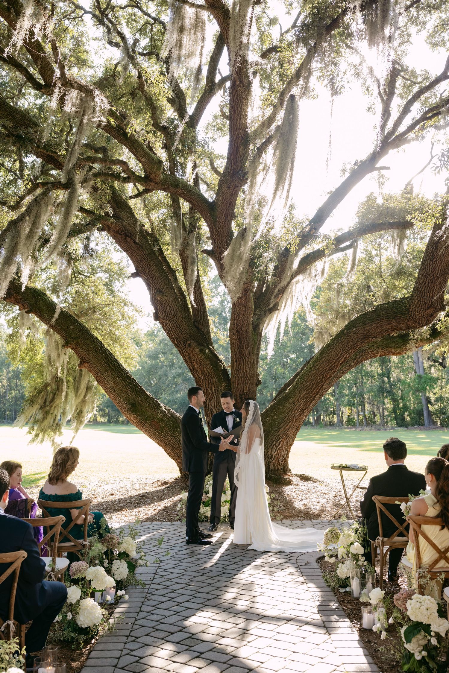 Romantic outdoor wedding ceremony setup under majestic oak trees draped with Spanish moss and decorated with white floral arrangements.