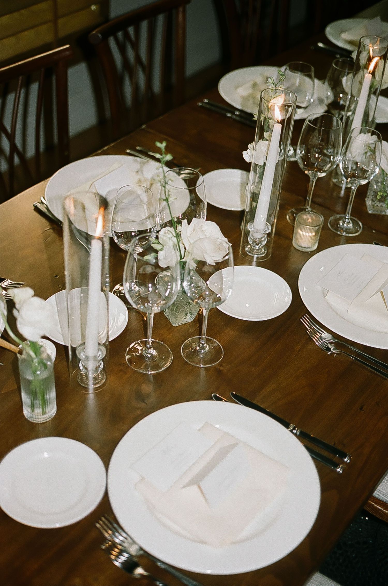 Place settings with white plates and wine glasses on wooden tables at a wedding reception.