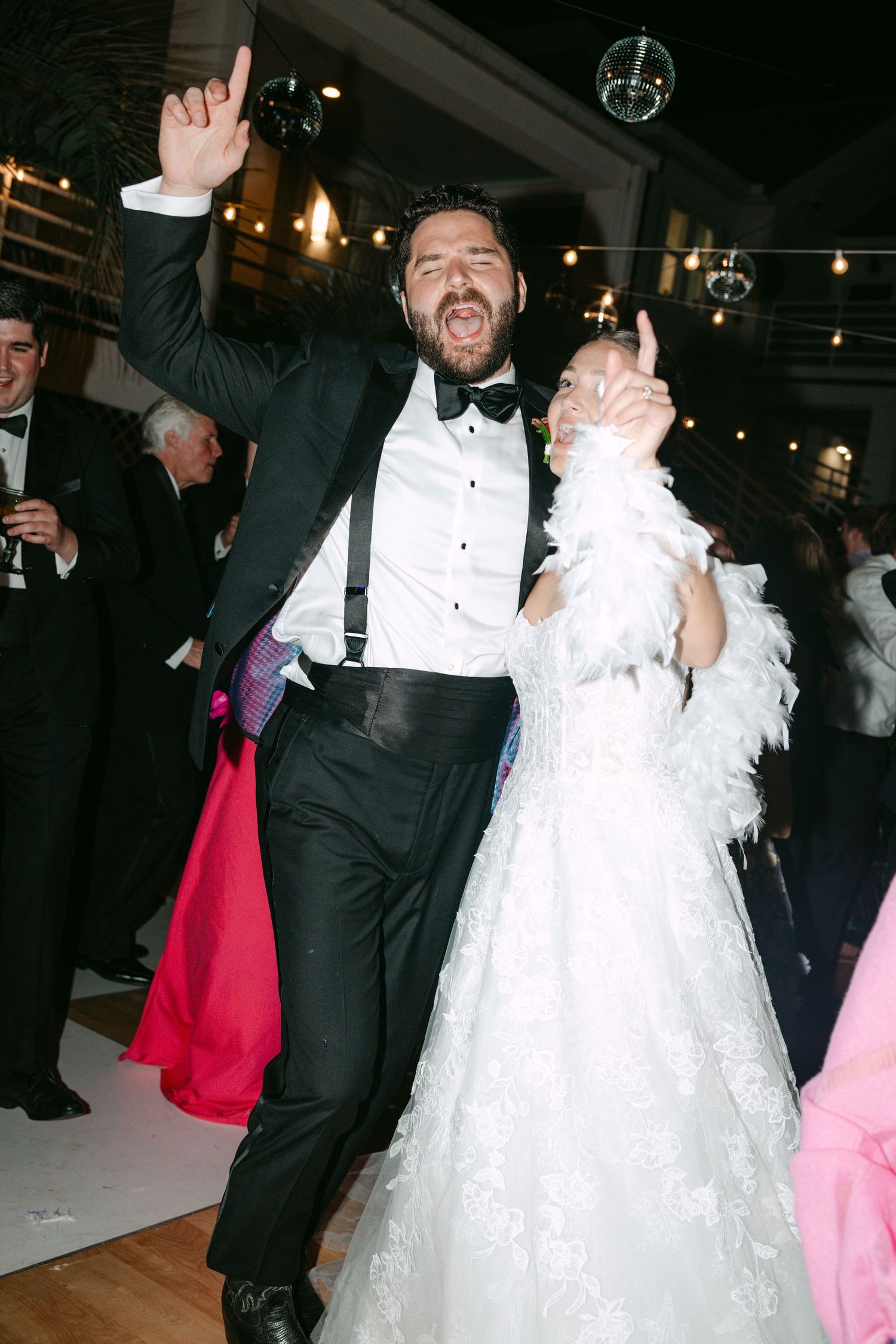 Joyful bride and groom dancing at wedding reception, bride wearing feather wrap over lace gown, groom in black tuxedo.