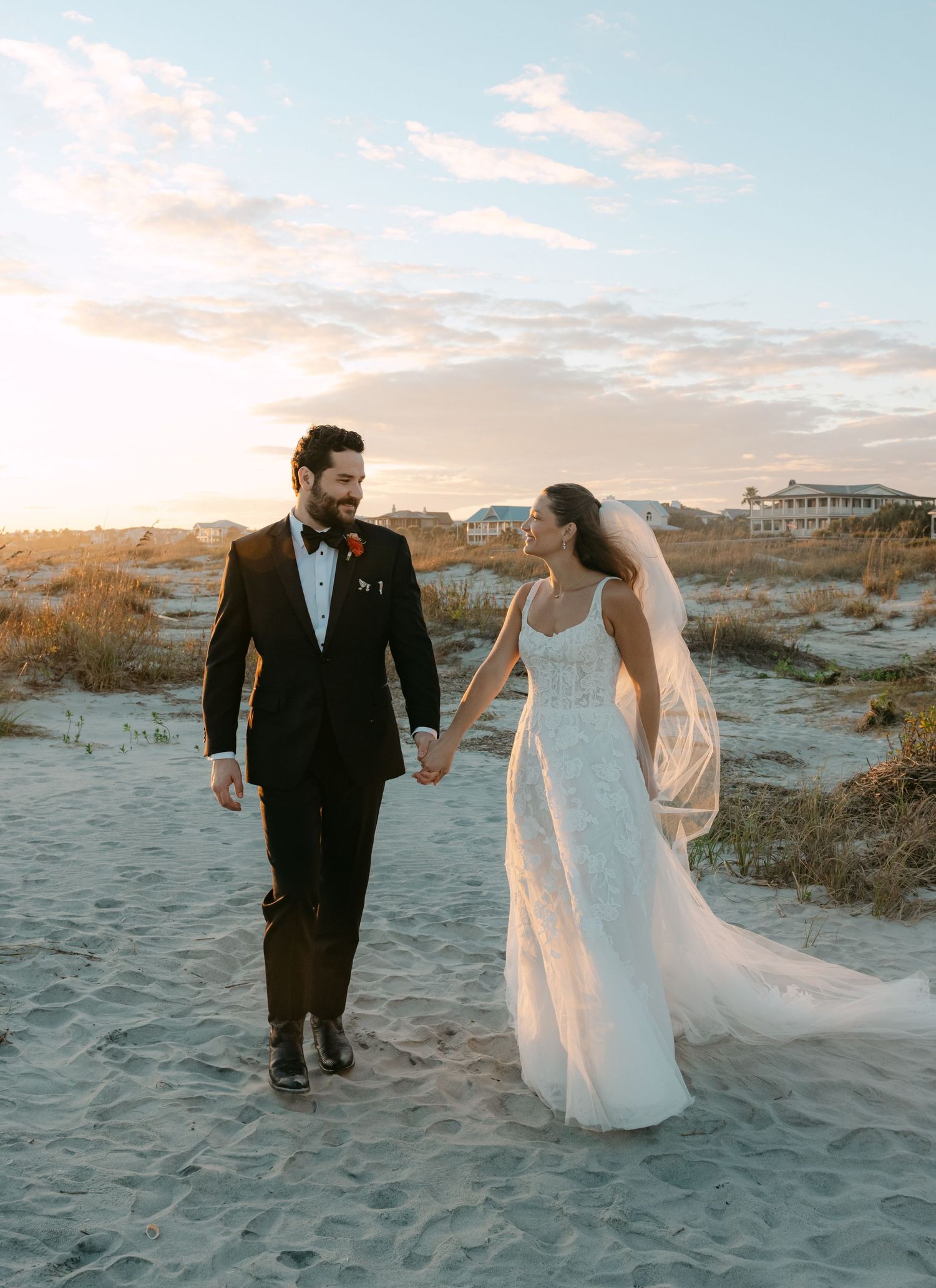 A bride and groom walk hand-in-hand on a sandy beach at sunset, she in a white gown with veil, he in a black tuxedo.