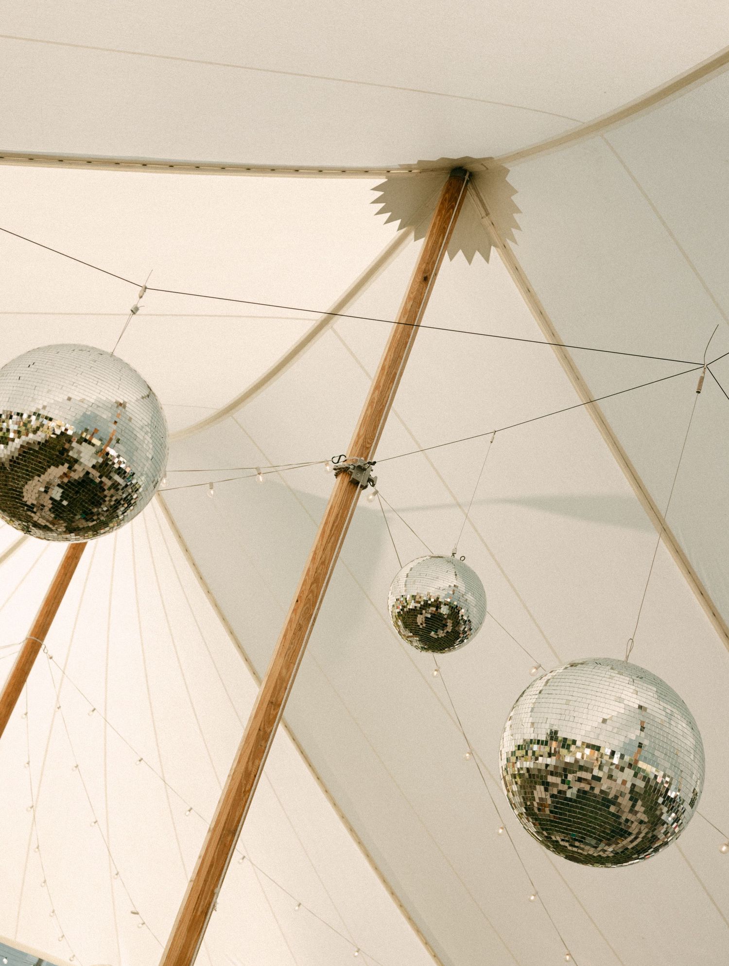 Disco balls hanging from the ceiling of a white sailcloth wedding tent with string lights.