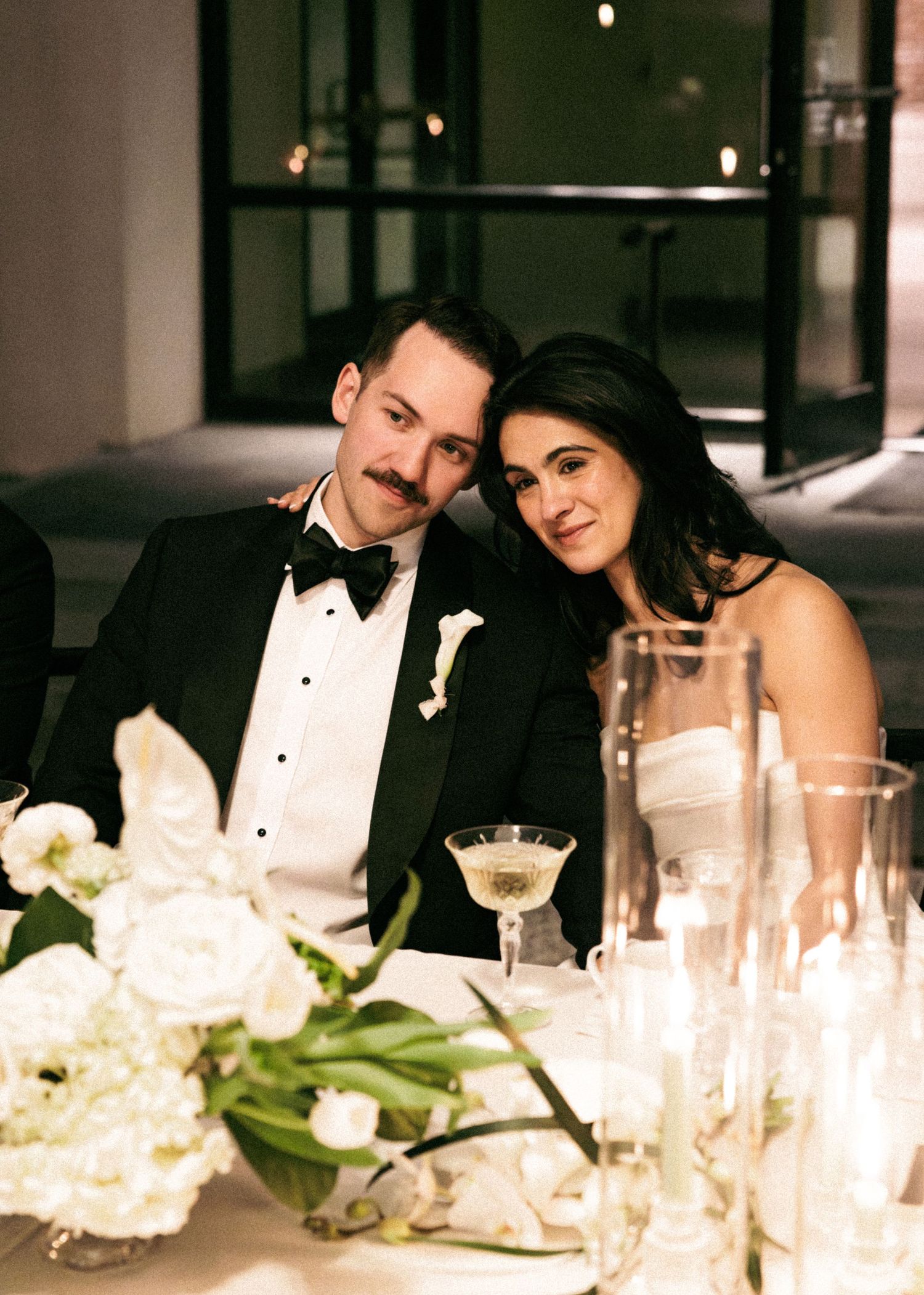 Couple in formal wedding attire sitting together at candlelit reception table with white floral arrangements.