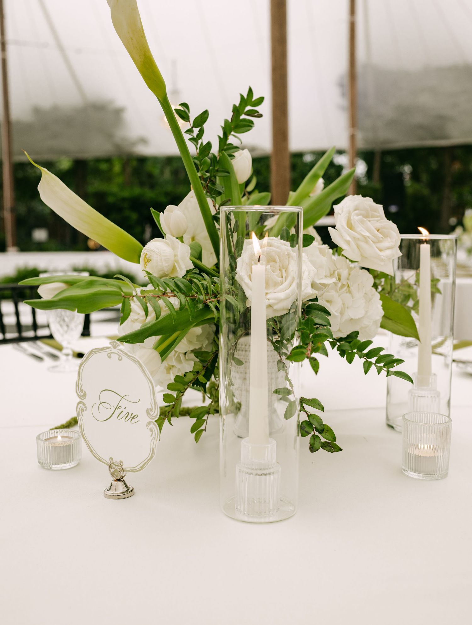 Elegant wedding table centerpiece with white roses, greenery, tall taper candles in glass holders, and a table number sign.
