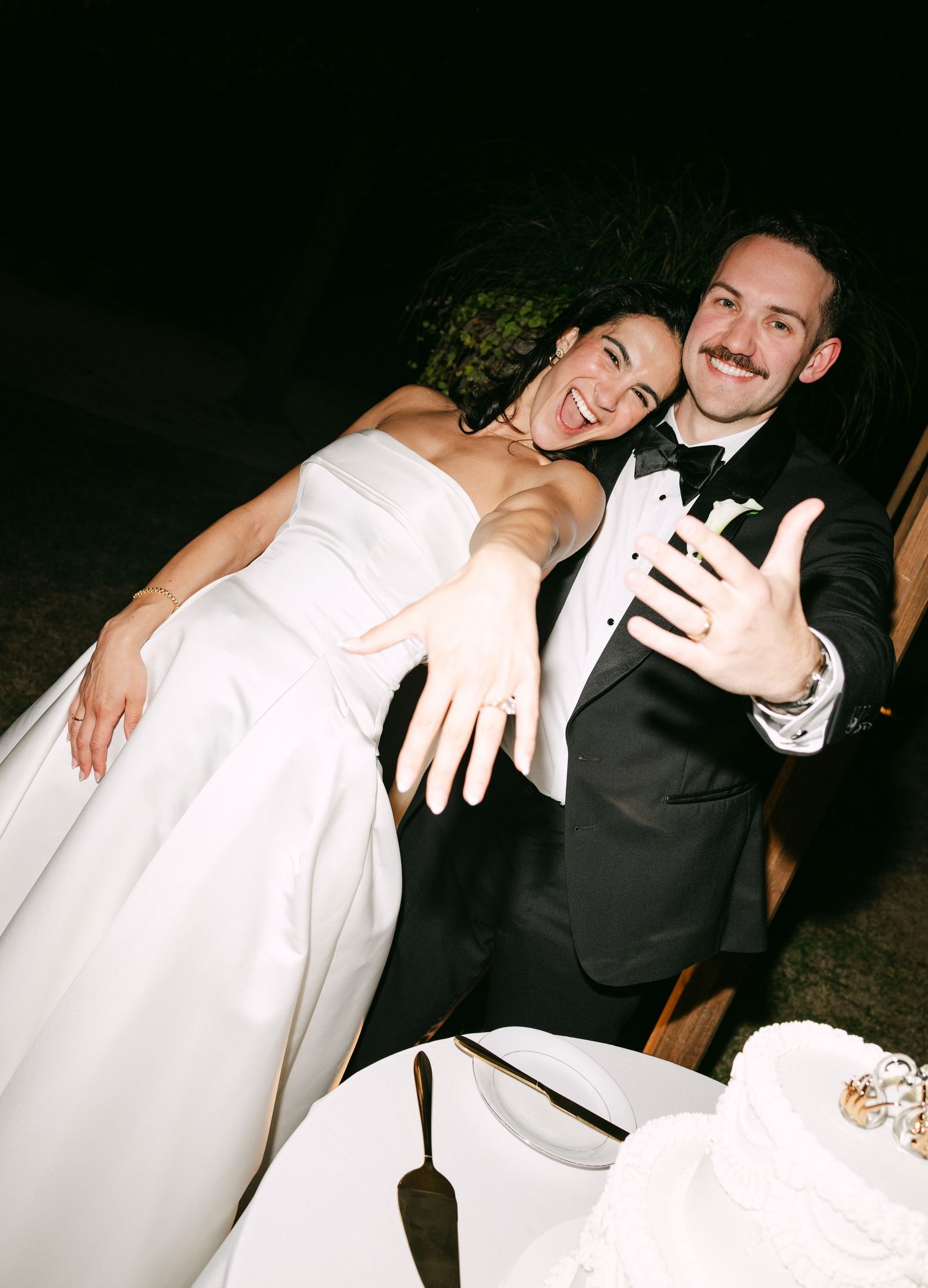 Smiling newlyweds showing their wedding rings beside a white tiered wedding cake.
