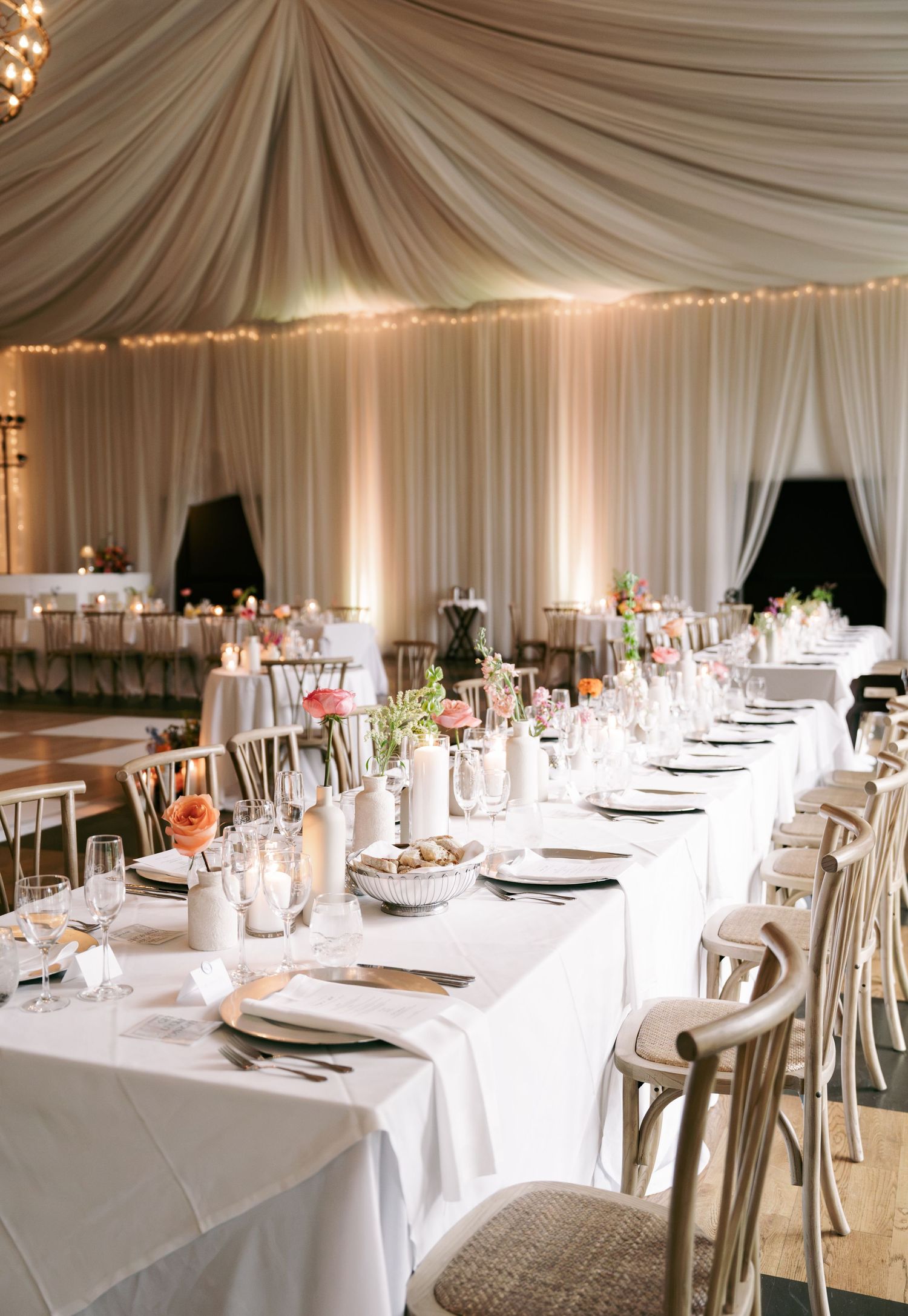 Elegant wedding reception hall with draped ivory ceiling, string lights, long tables and pink floral centerpieces.