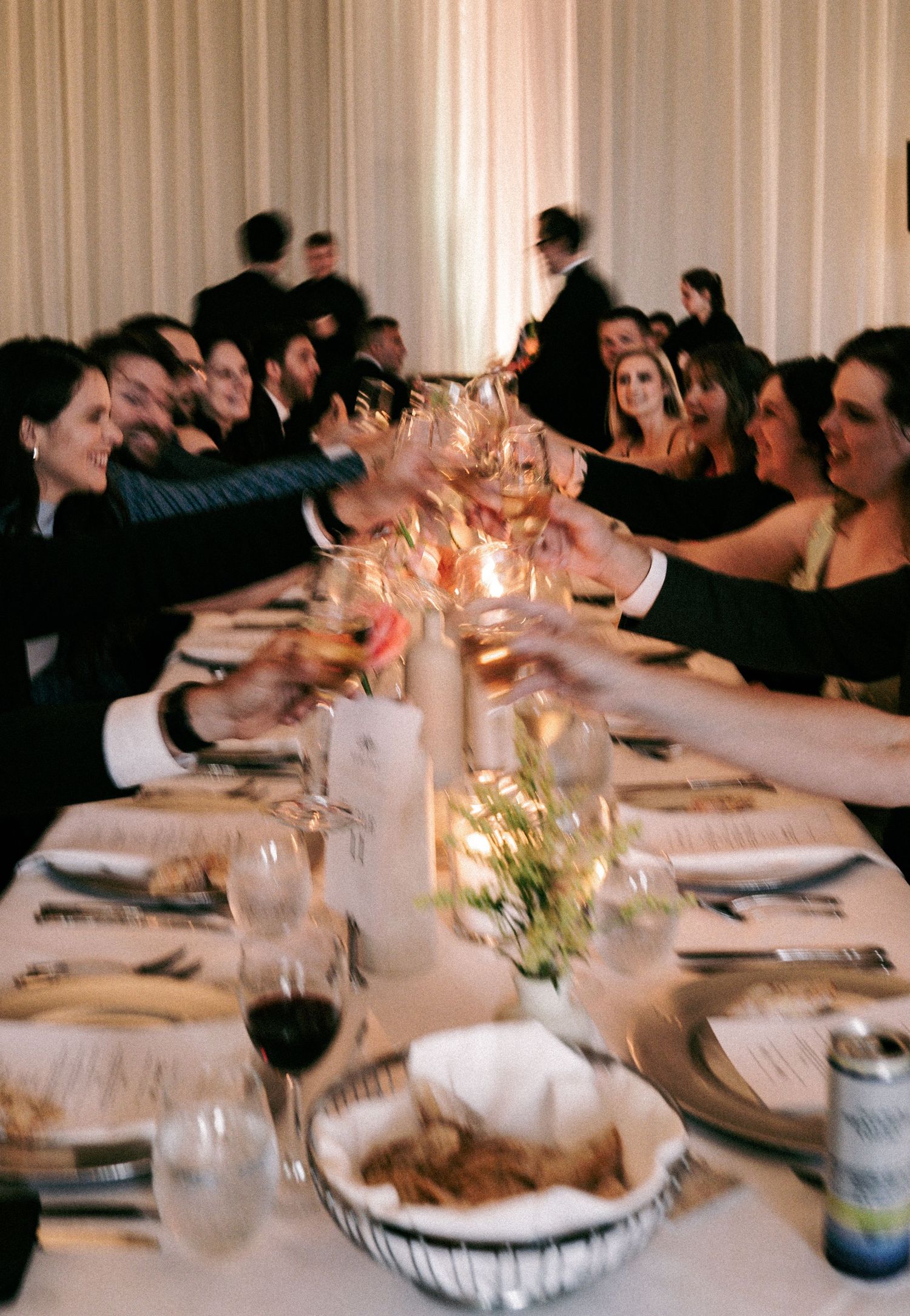 Wedding guests raising glasses in a toast at a long reception table with candlelight and white drapes.