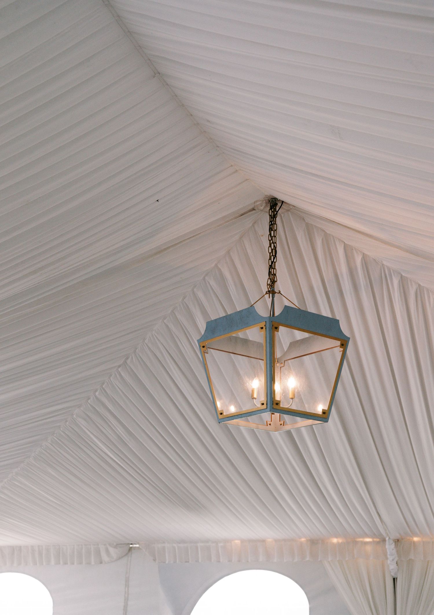 Elegant brass lantern chandelier hanging from draped white fabric ceiling inside a wedding reception tent.