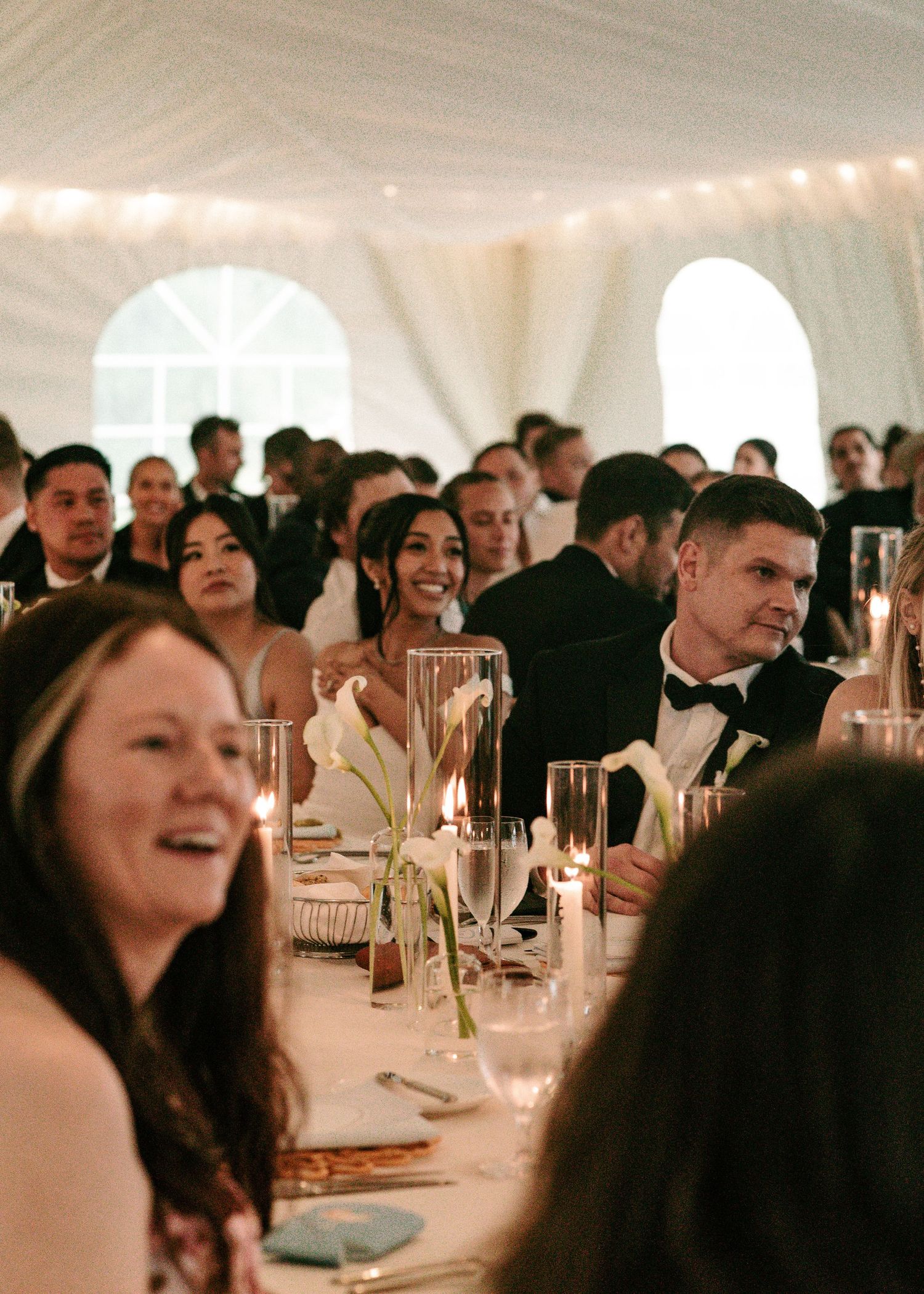 Bride and groom smile at guests during candlelit wedding reception inside an elegant white draped tent.