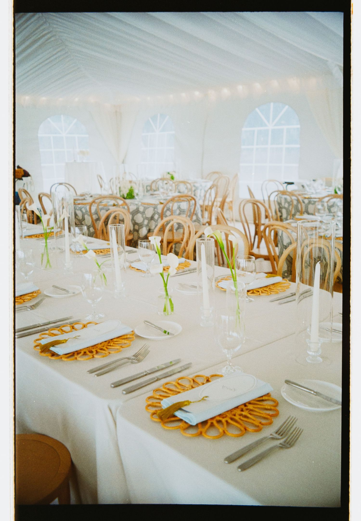 Elegant white tent reception with rattan chairs, yellow charger plates, taper candles, and white floral centerpieces on long tables.