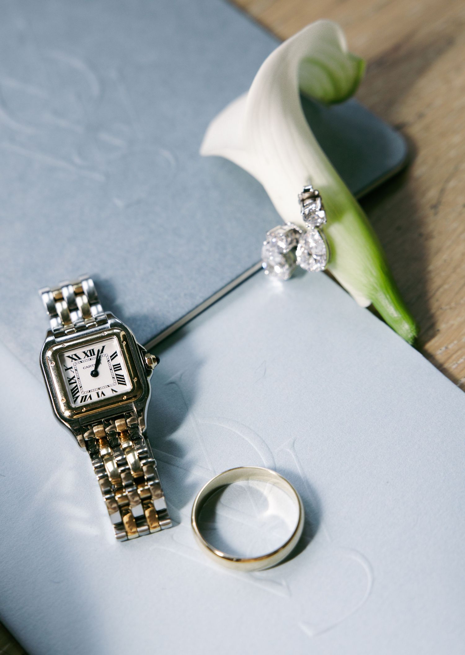 Cartier watch, gold wedding band, diamond earrings, and white calla lily arranged on a blue embossed book.