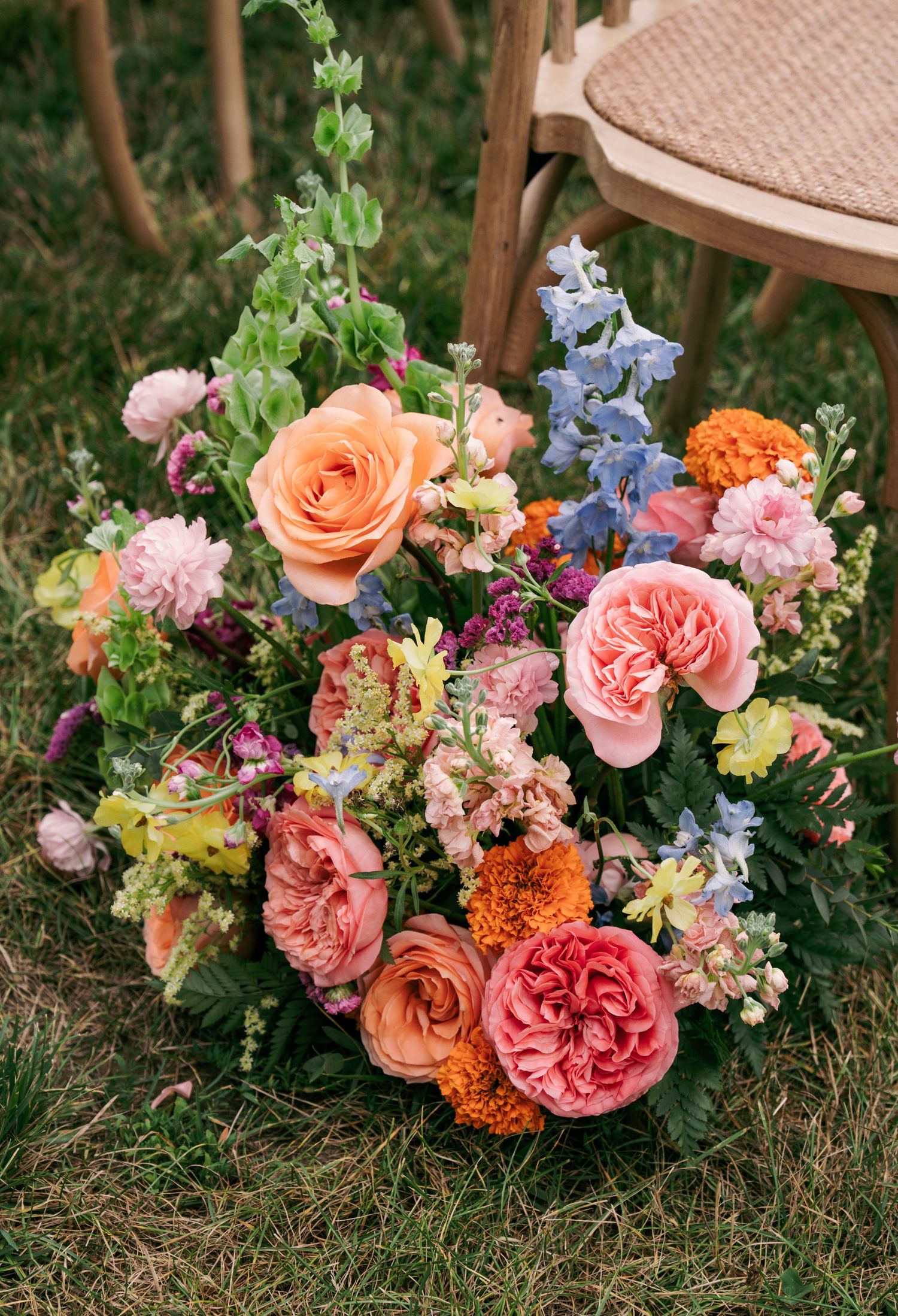 Colorful wedding bouquet with peach roses, pink blooms, blue delphiniums, and orange flowers on grass.