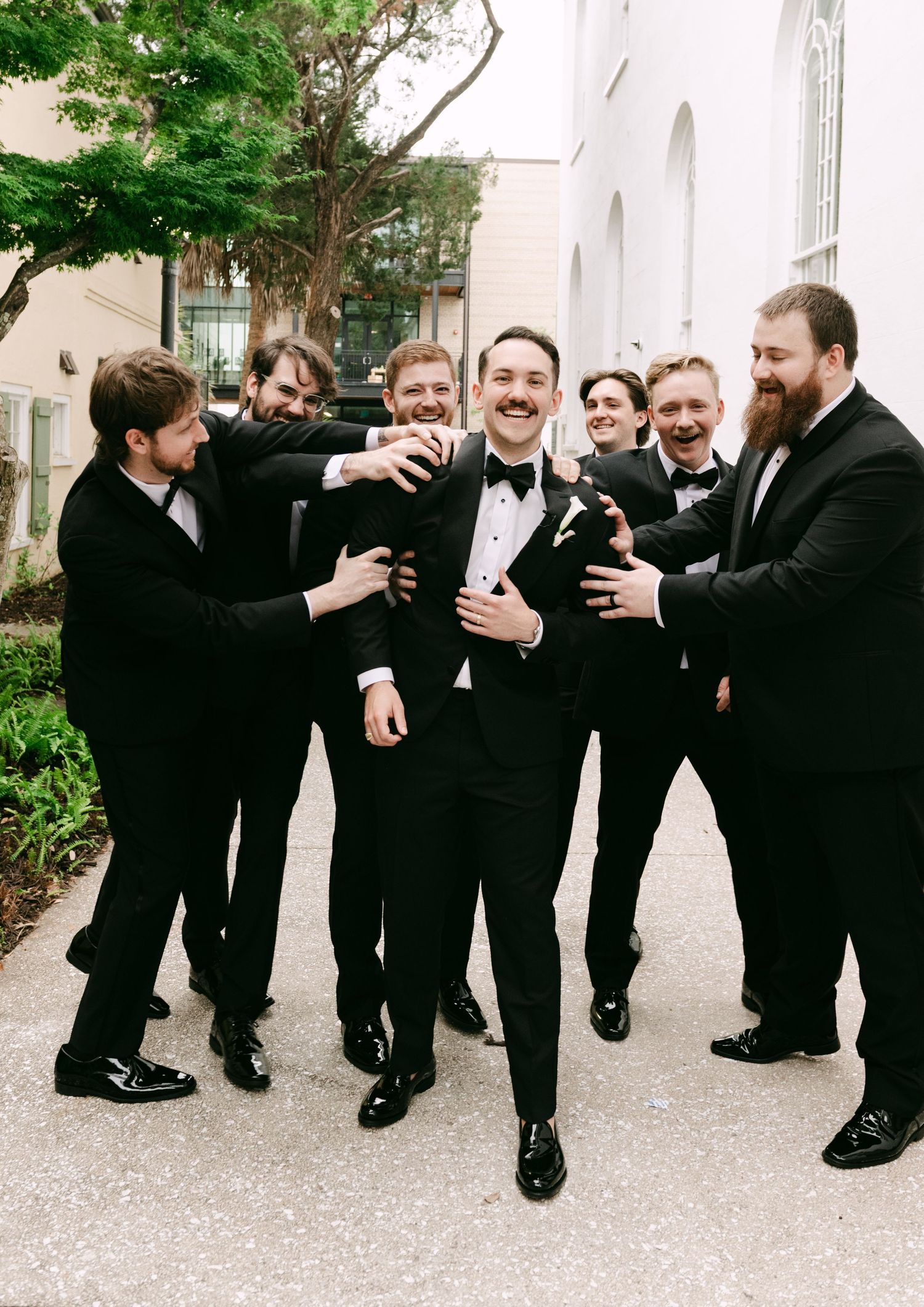 Groom in black tuxedo laughing with groomsmen in matching suits at outdoor wedding venue.