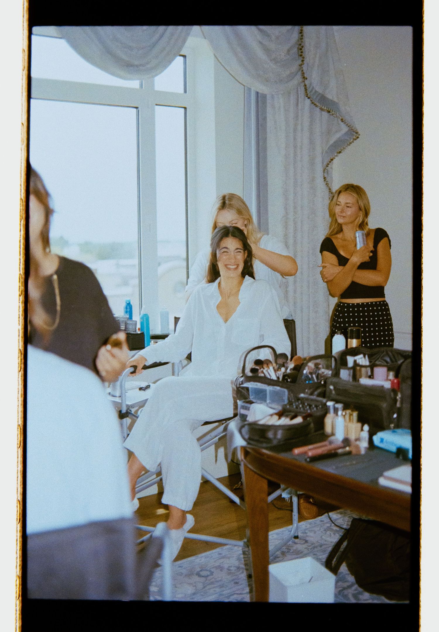 Bride in white pajamas getting hair styled by bridesmaids in bright hotel suite before wedding.