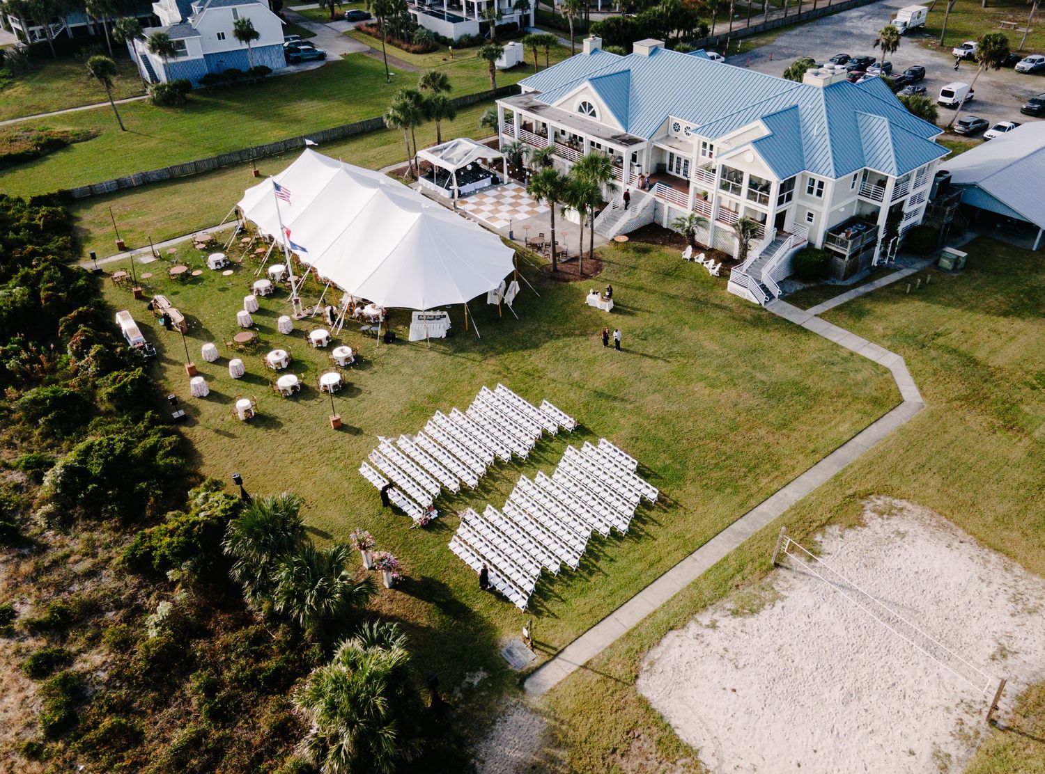 Aerial view of outdoor beach wedding venue with white tent, ceremony chairs, and coastal mansion.