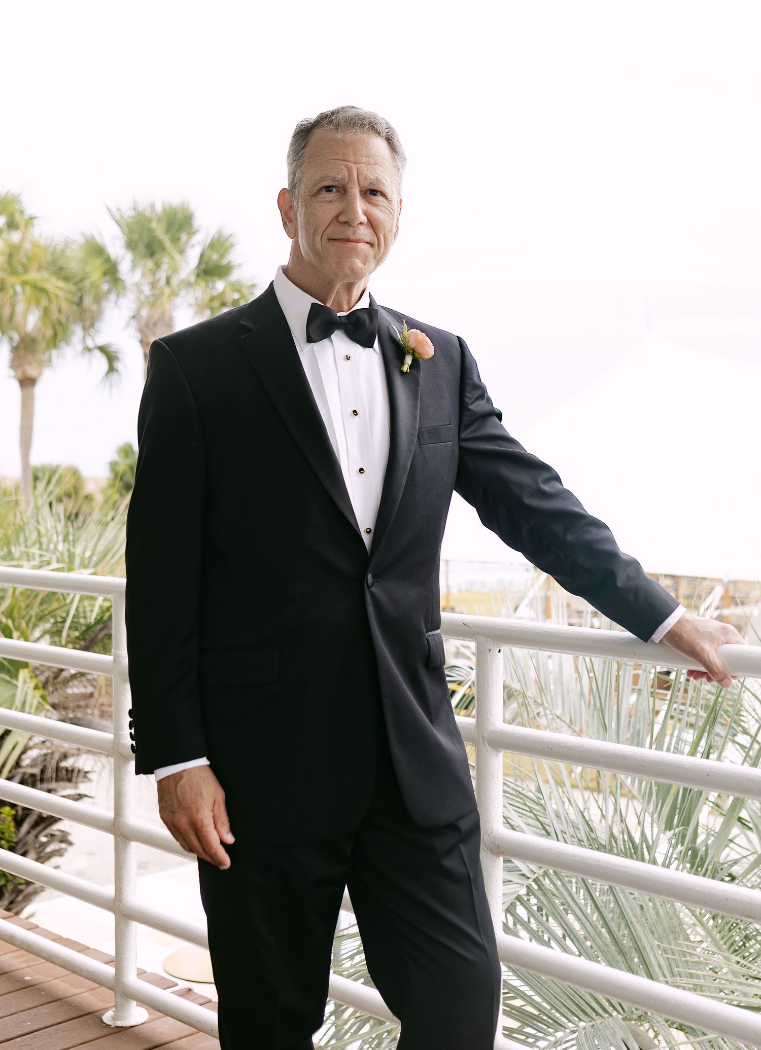 Elegant groom in black tuxedo with bow tie and boutonniere poses on waterfront deck with palm trees.