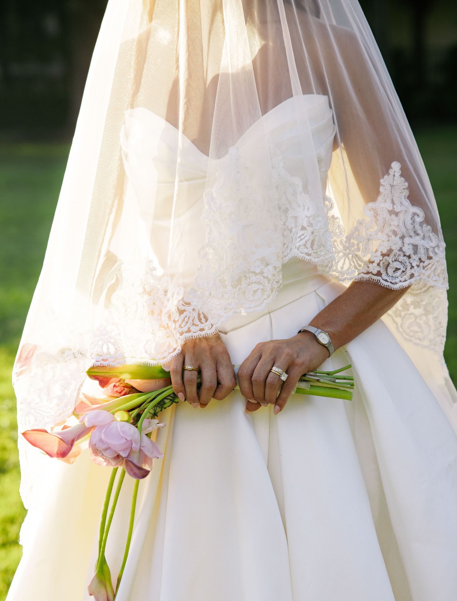 Bride in lace wedding gown and veil holding pink tulip bouquet, showcasing diamond engagement ring and bracelet.