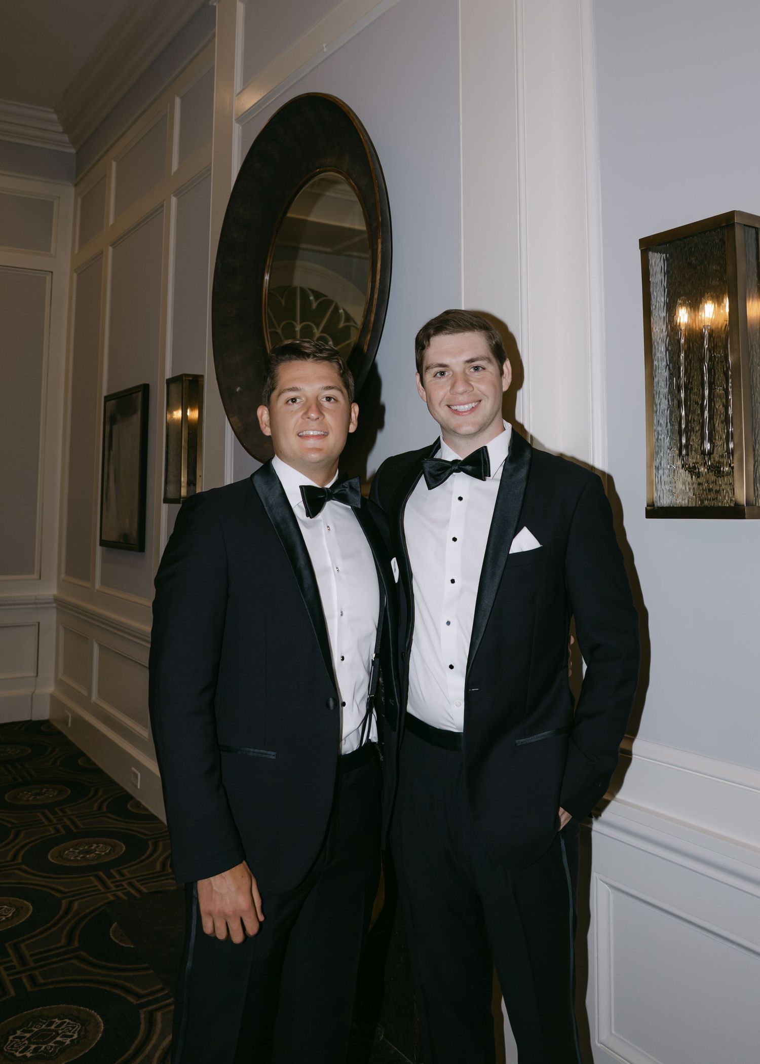 Two people in matching black tuxedos pose in elegant hallway.