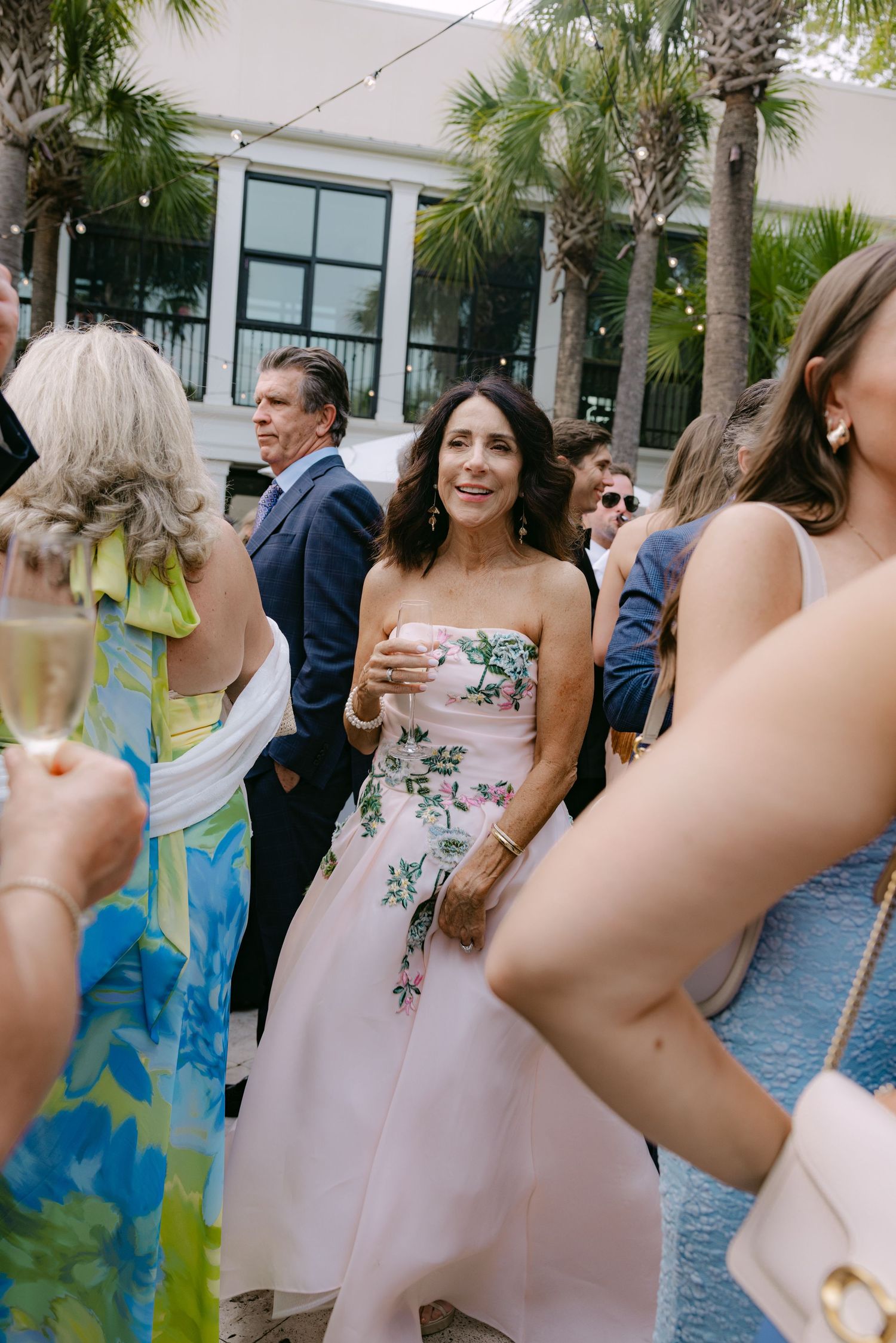 Guests in floral dresses enjoying an outdoor reception under palm trees.