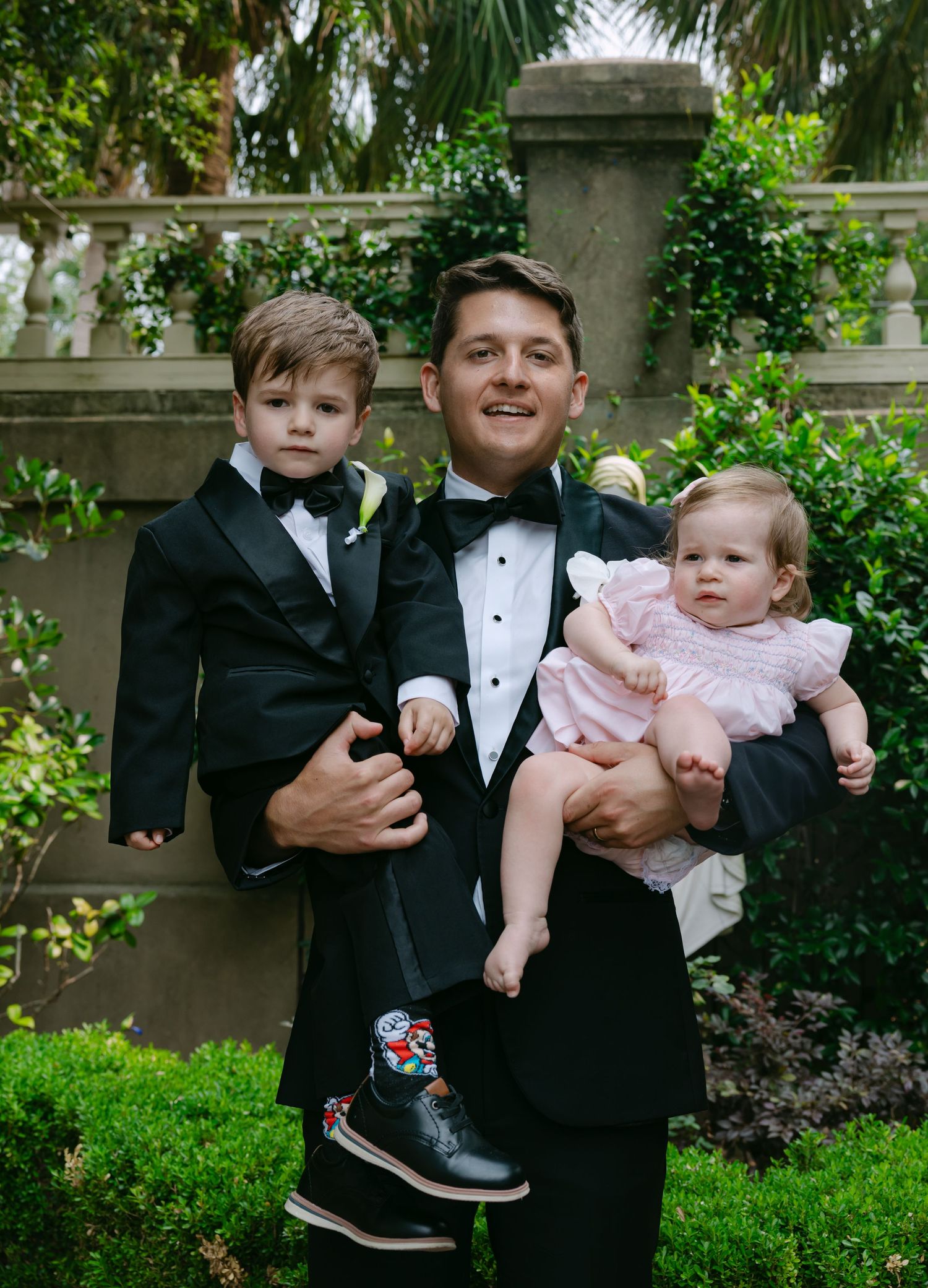 Young children in formal wedding attire being held by adults in garden setting.