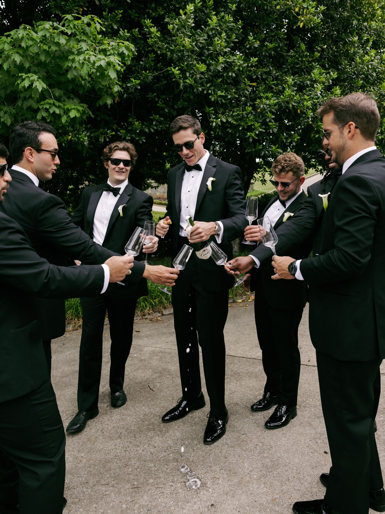 Groomsmen in black tuxedos toasting with champagne outdoors, celebrating before a wedding ceremony.
