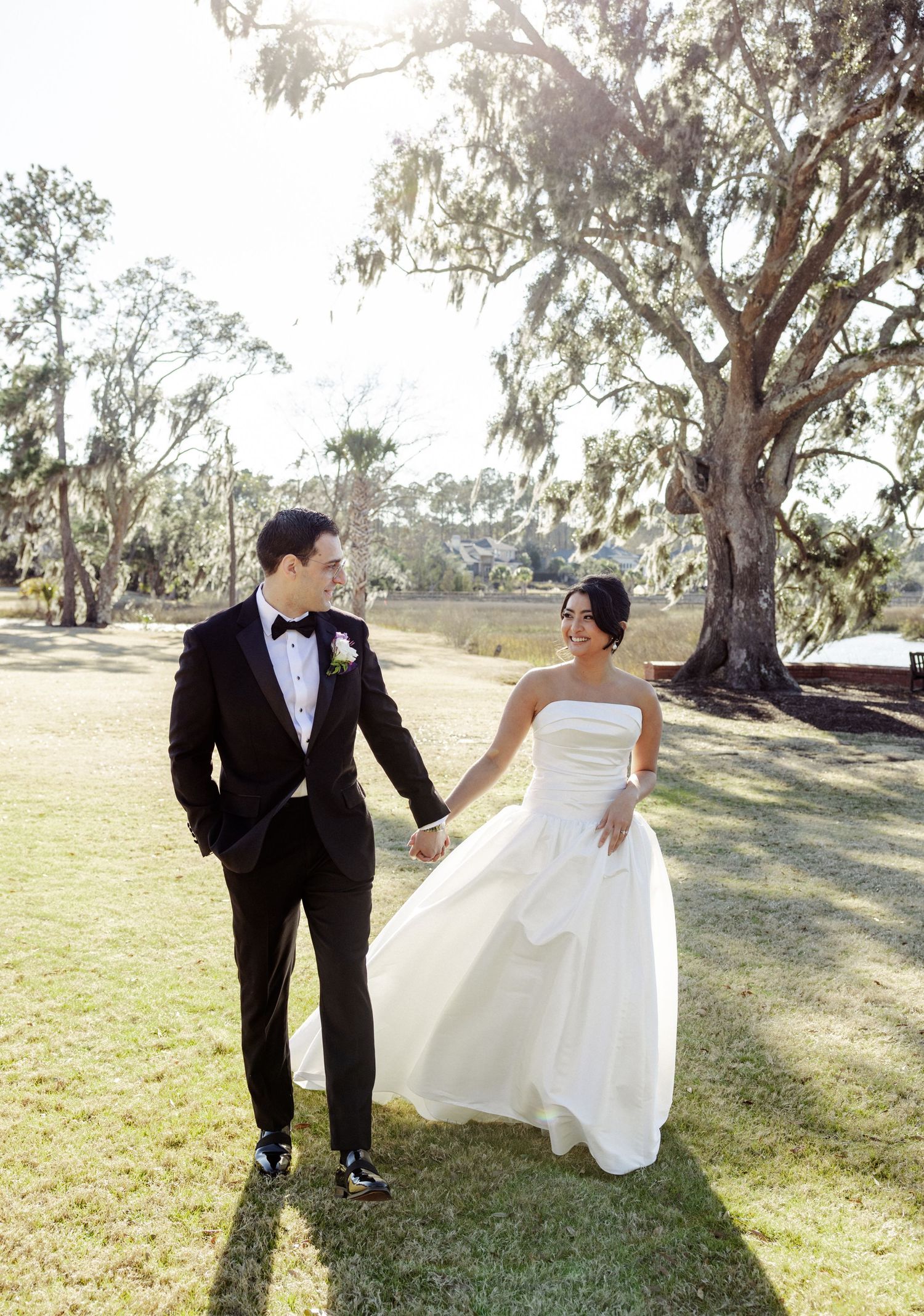 A couple in wedding attire holds hands while walking through a sunlit park with large eucalyptus trees.