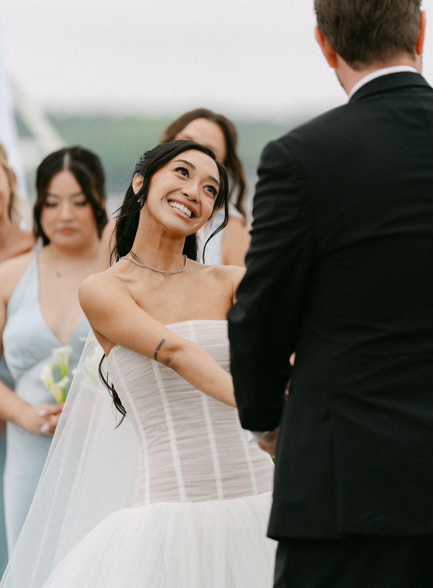 Bride in strapless white gown smiling joyfully at groom during outdoor wedding ceremony with bridesmaids in blue.