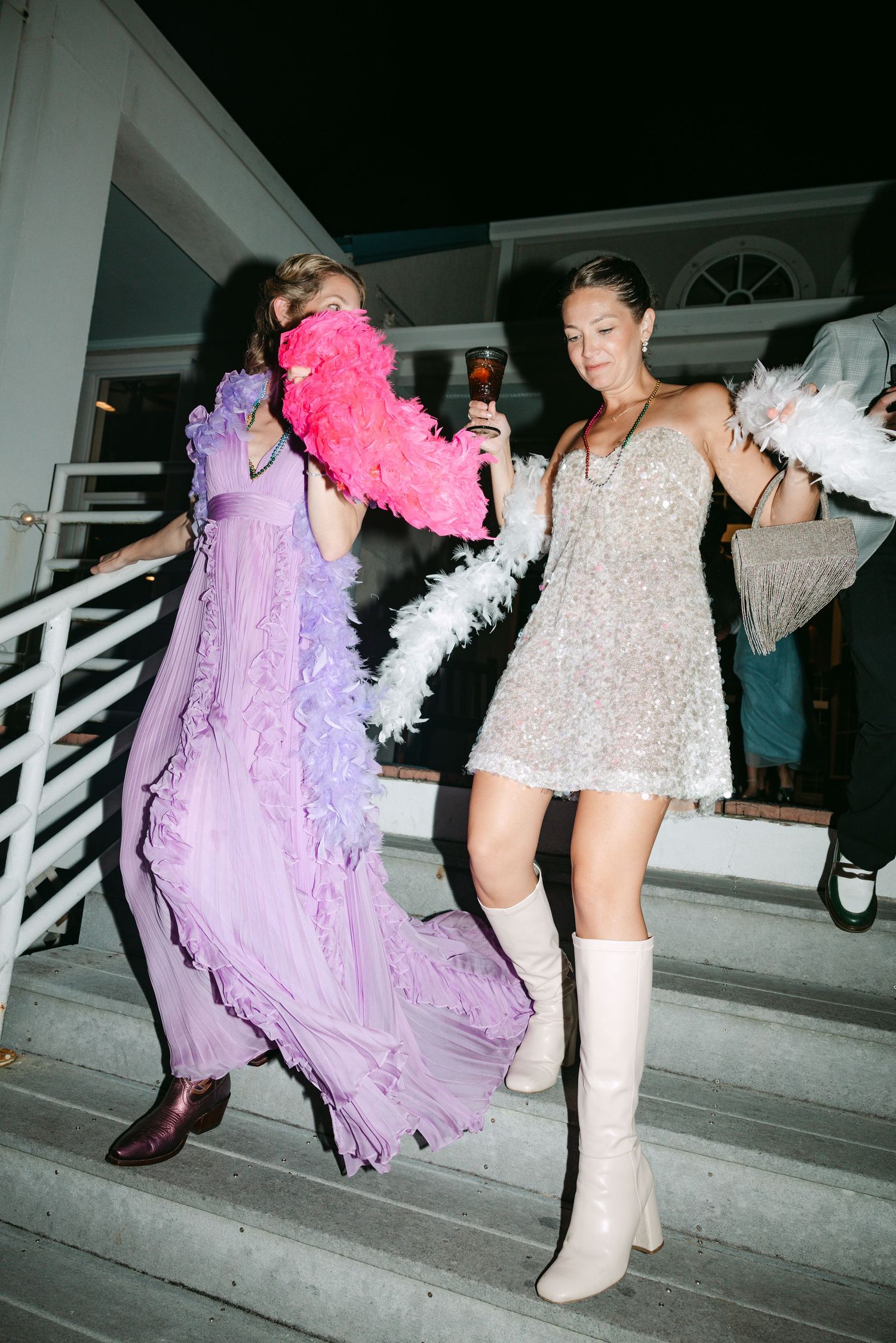 Two women in festive dresses with feather boas dancing on outdoor stairs at night.