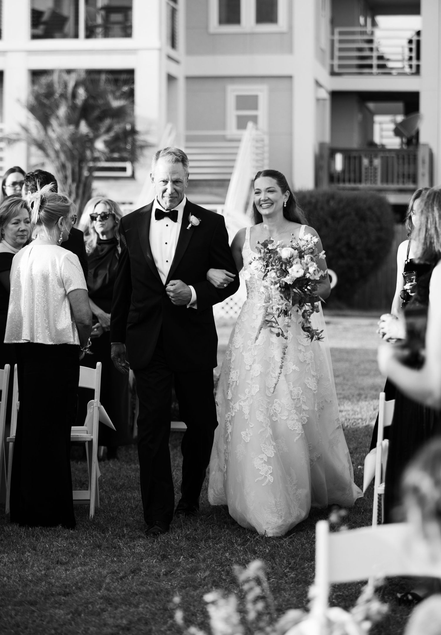 Black and white photo of bride walking down outdoor aisle with father in tuxedo at wedding ceremony.