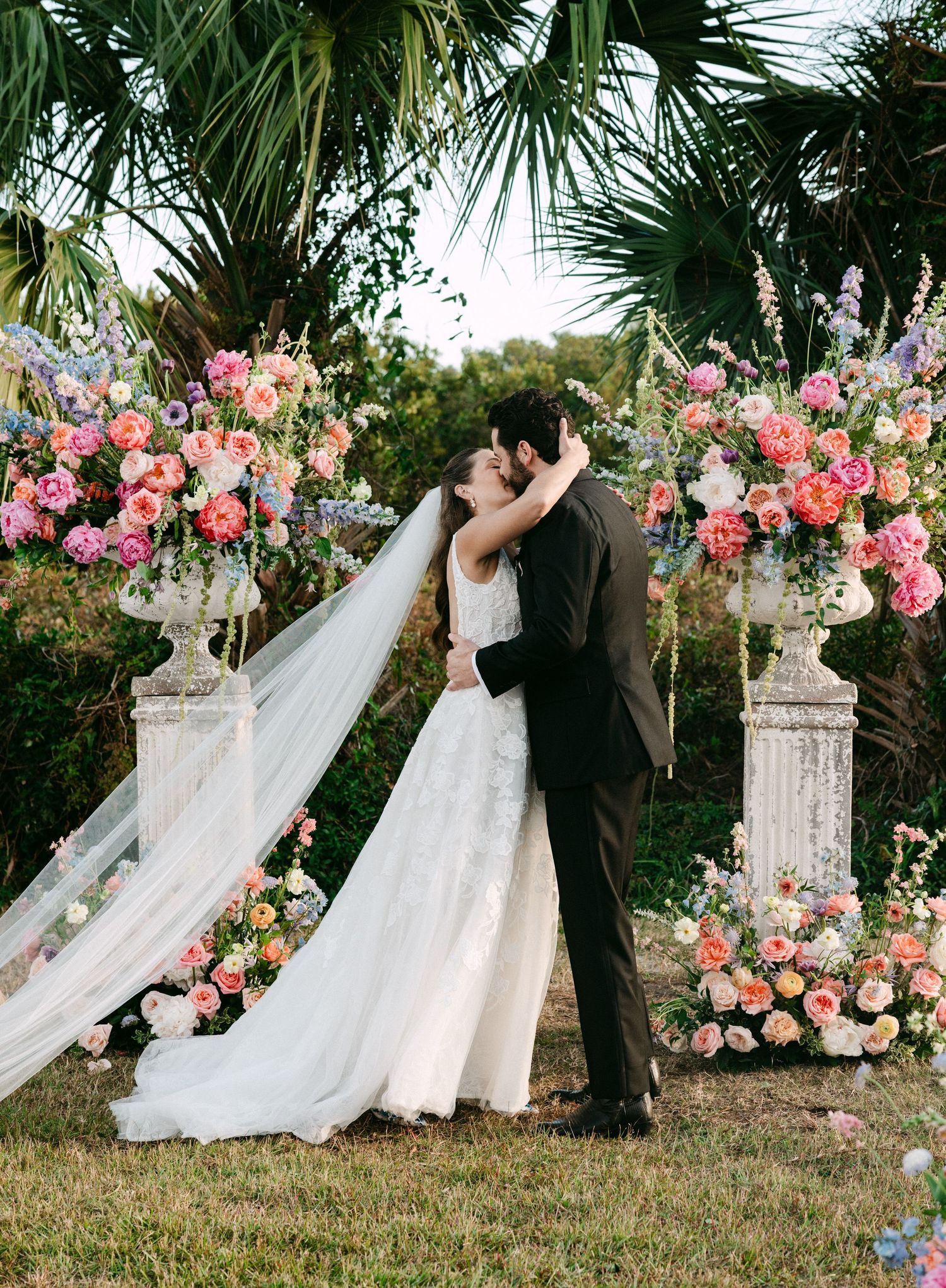 Bride and groom share first kiss at outdoor altar flanked by colorful floral arrangements on pillars.