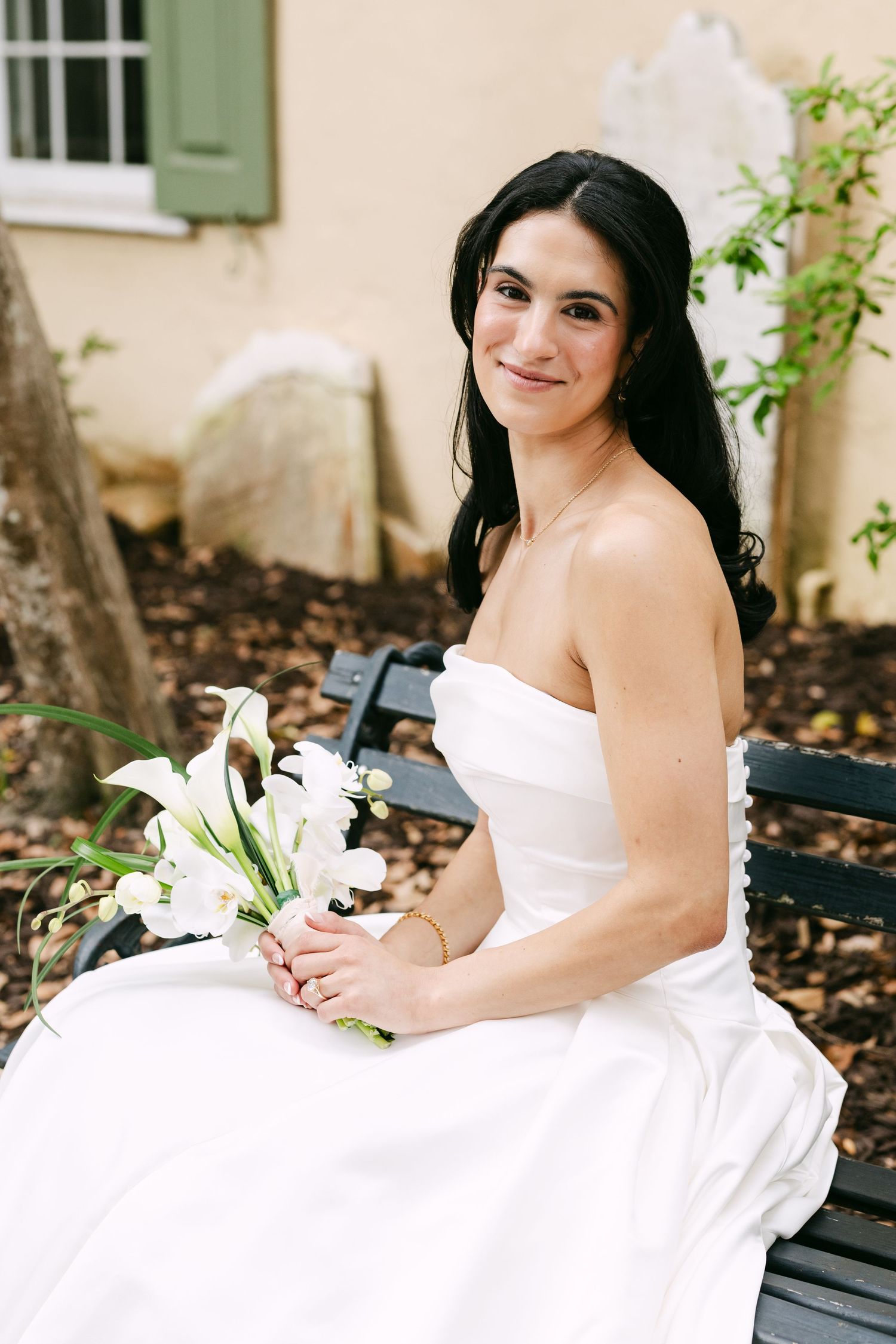 Bride in strapless white gown holding white calla lily bouquet while seated on a black bench in a garden.