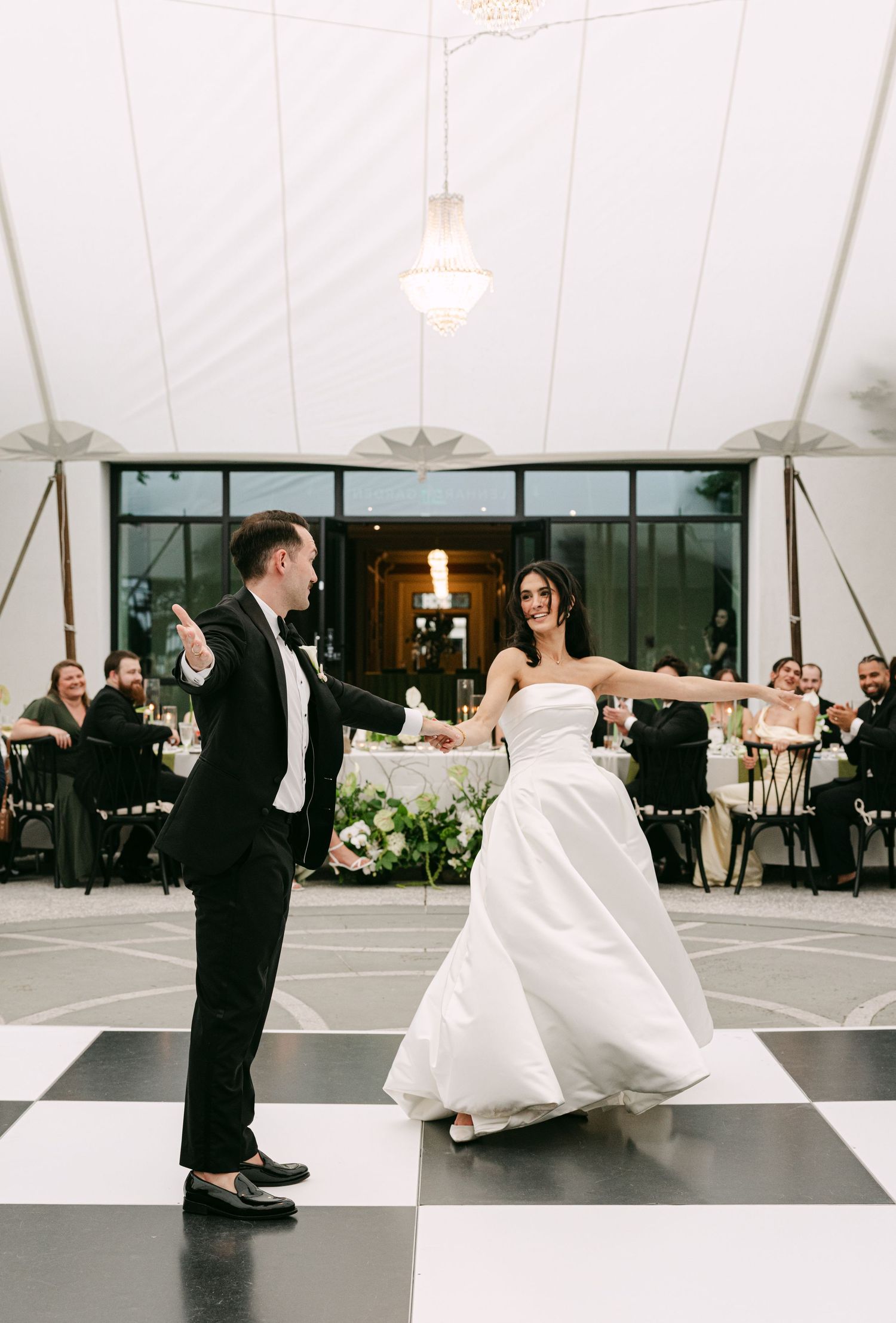Bride and groom first dance on black and white checkered floor under elegant tent with crystal chandelier.