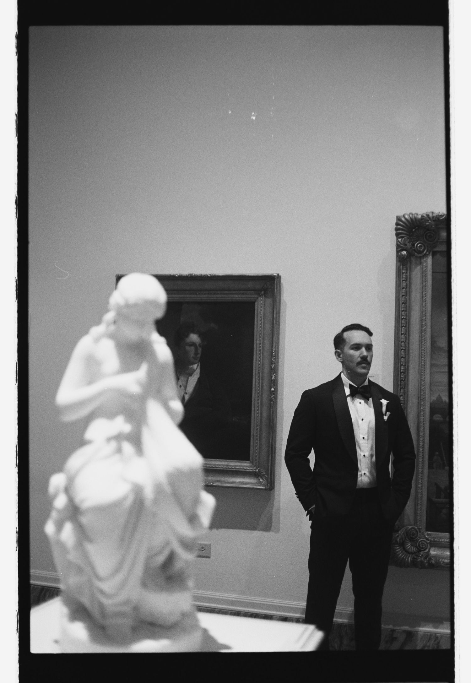 Groom in tuxedo standing beside classical white marble sculpture in black and white museum photo.