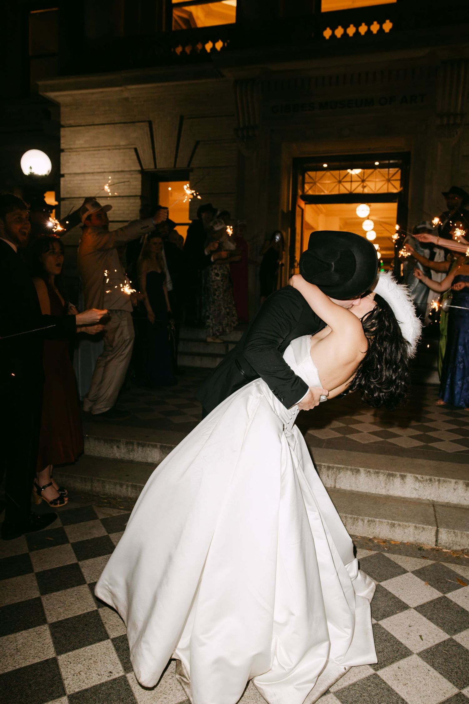 Bride and groom share romantic dip kiss on checkered steps surrounded by sparkler-holding wedding guests at night.