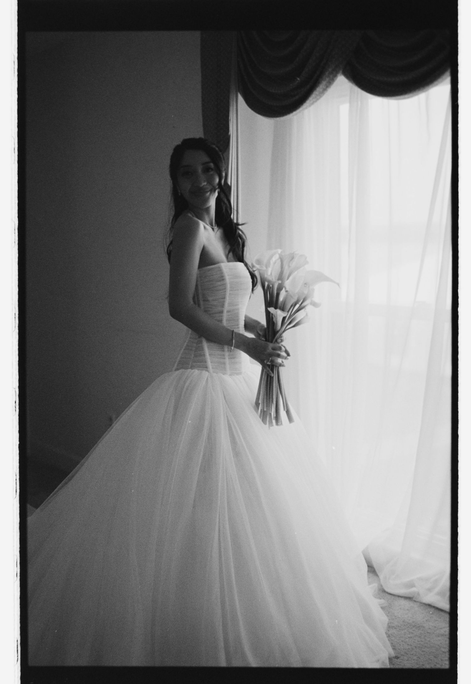 Black and white photo of a smiling bride in a tulle gown holding calla lilies near a draped window.