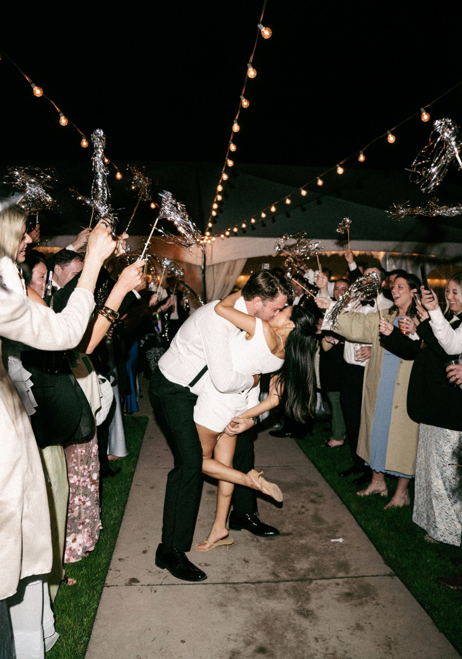 Groom dips bride in romantic wedding exit with sparklers and string lights at night outdoor reception.